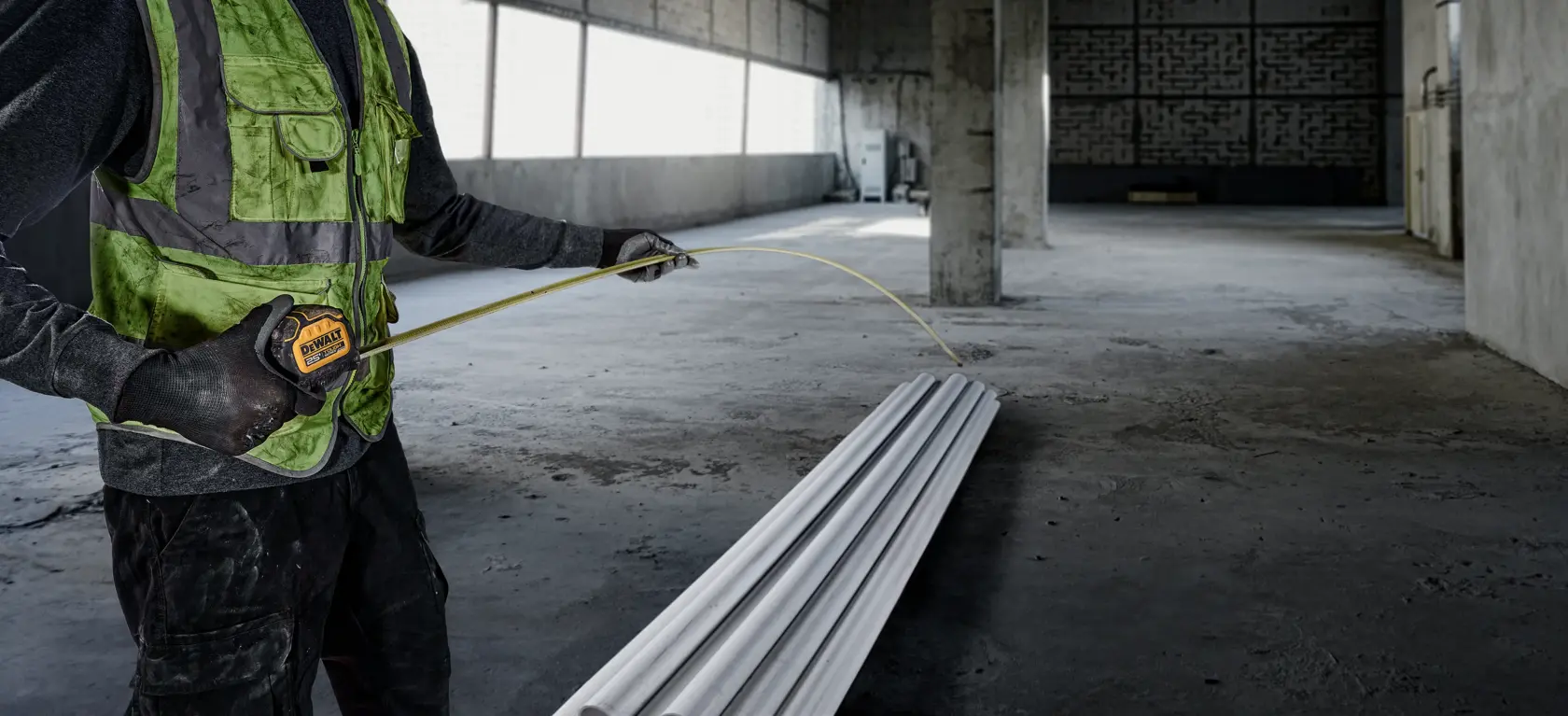 A person wearing a green safety vest and black gloves uses a DEWALT tape measure to measure metal pipes in an unfinished industrial building.