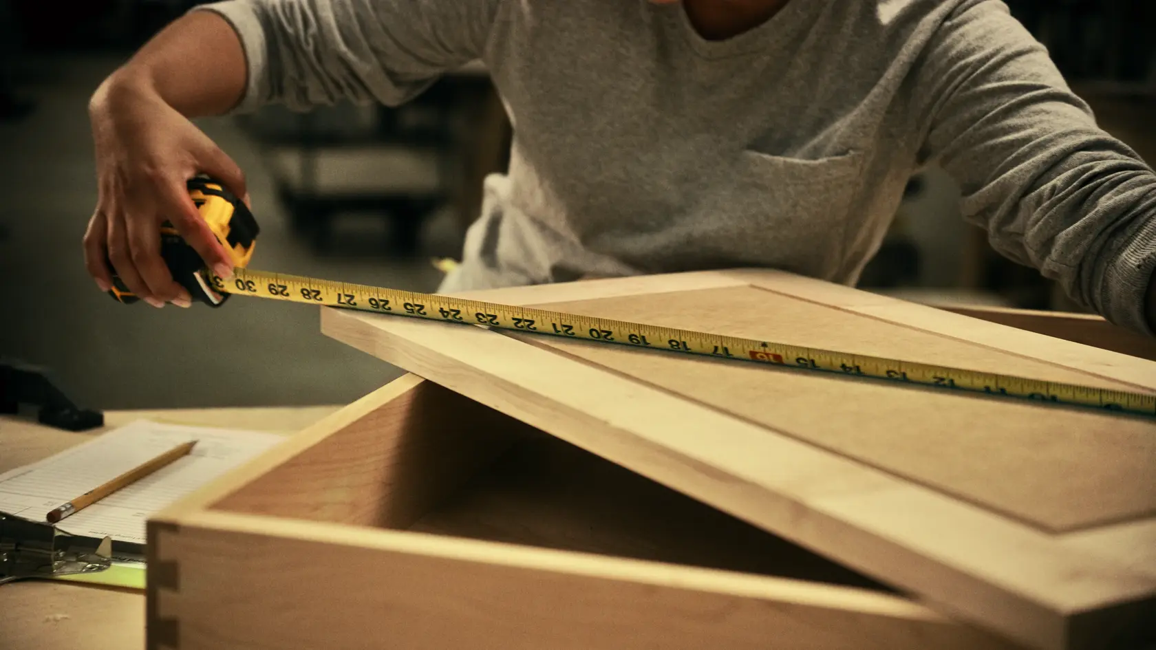 A person uses a DEWALT tape measure to measure the length of a wooden board or box lid on a workbench.