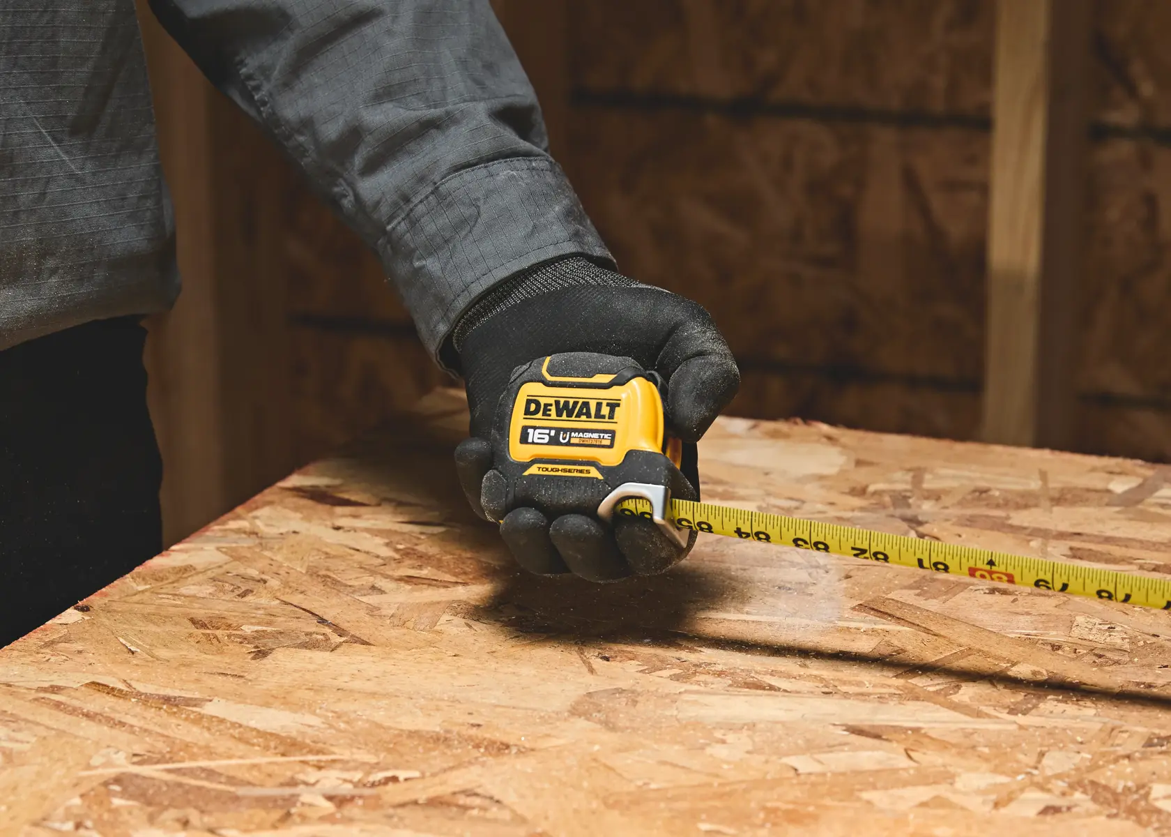 A person wearing a black glove is holding a DEWALT 16-foot tape measure and measuring a wooden surface in a workshop or construction setting.