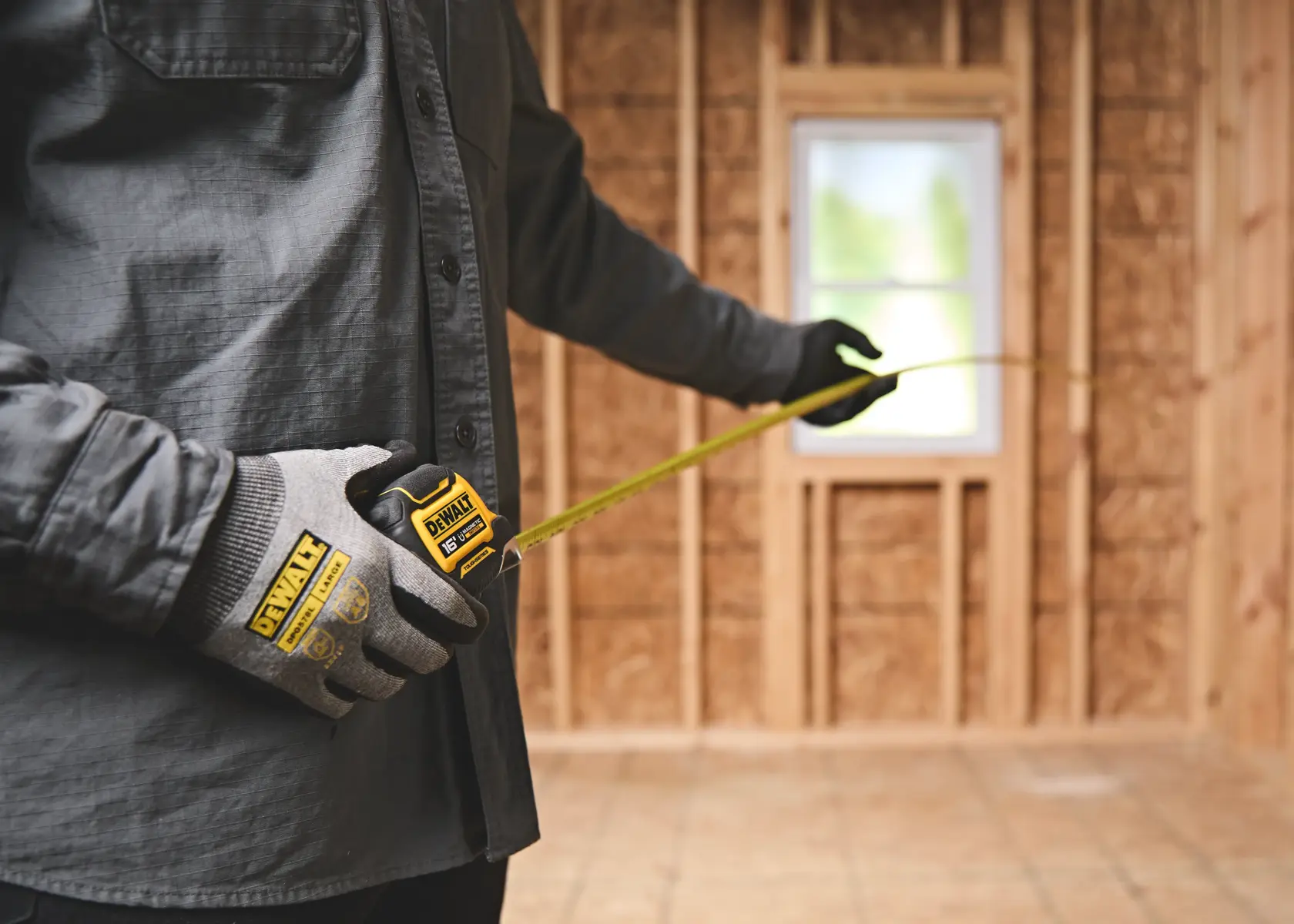 Person wearing DEWALT branded work gloves using a DEWALT tape measure in a wooden framed room under construction.