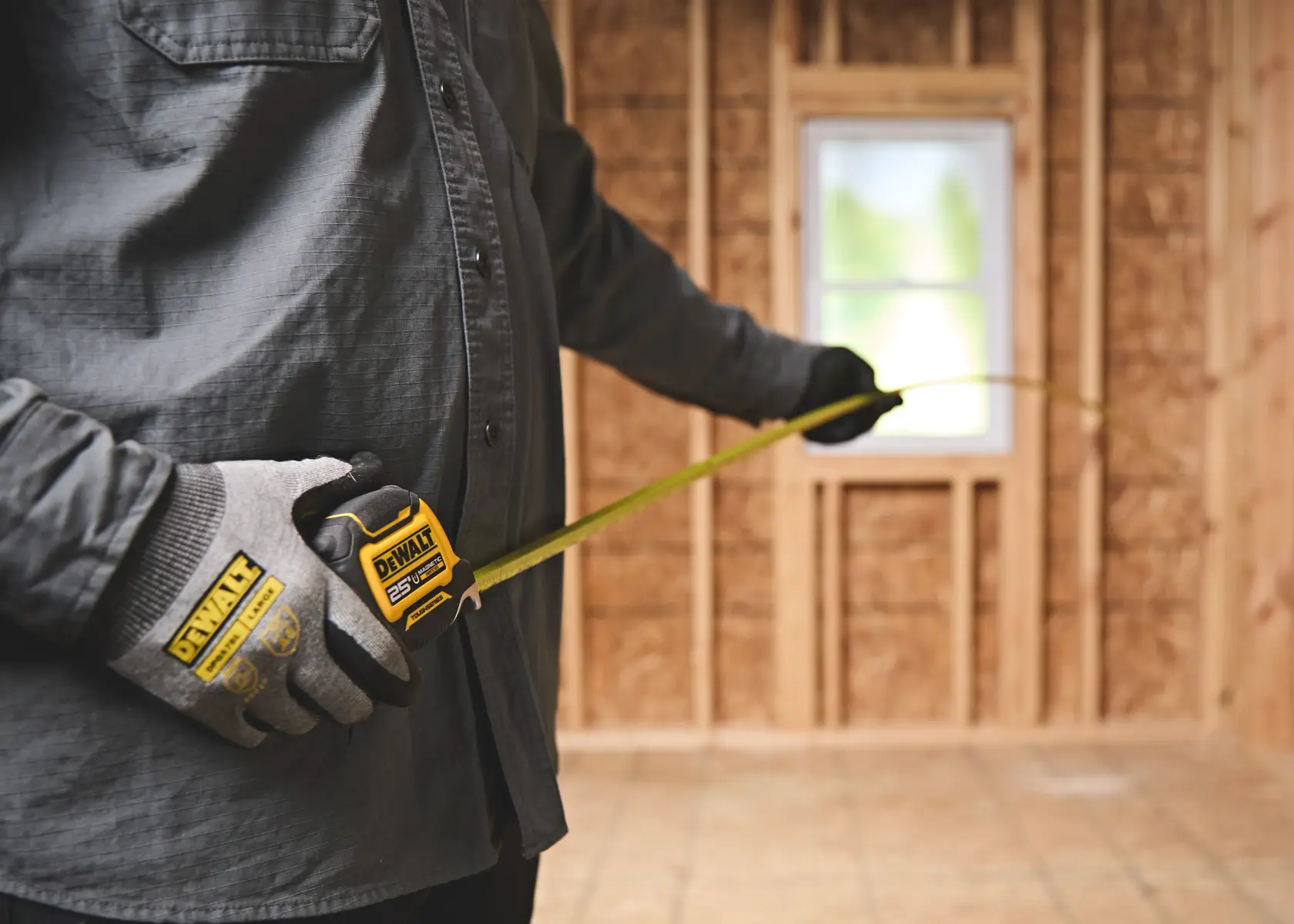 A person wearing gloves is using a DEWALT 25-foot tape measure in a room with unfinished wooden walls and a window.