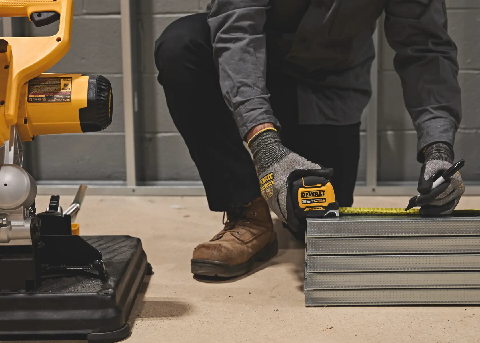 A person wearing gloves and work boots is using a DEWALT tape measure to mark metal studs, with a saw visible on the left side of the image in a workshop setting.