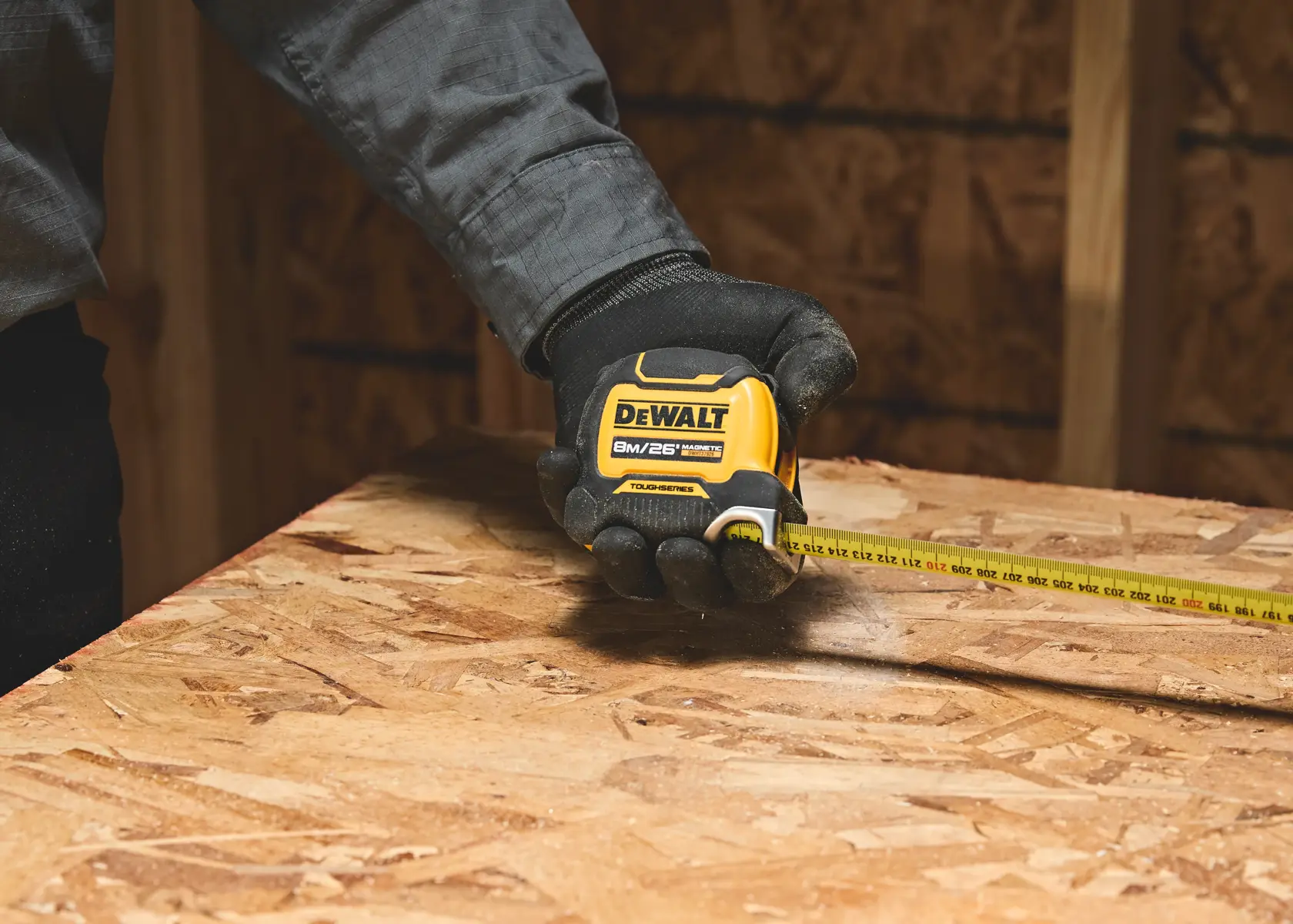 A person wearing a glove is using a DEWALT 8m/26ft tape measure to measure a piece of plywood in a construction setting.