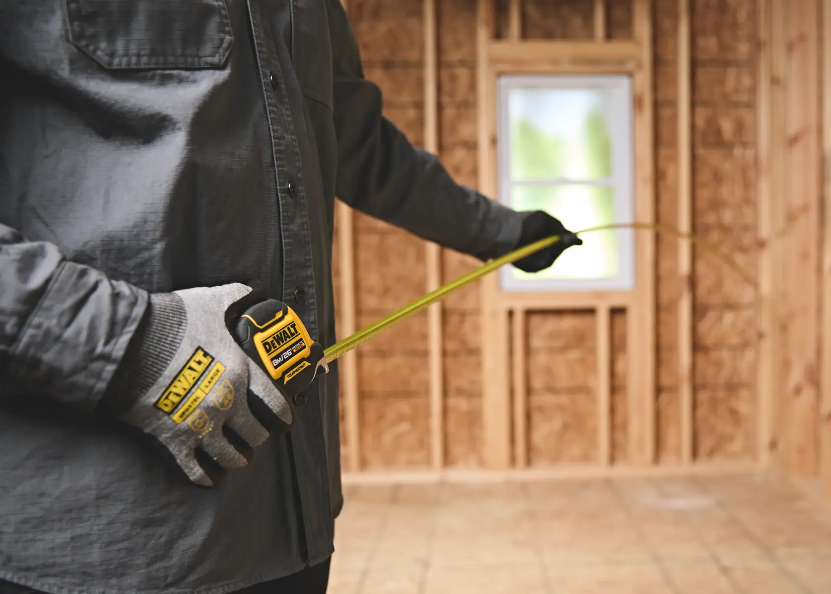 A person wearing work gloves is using a DEWALT tape measure in a room under construction with exposed wooden framing and a window.