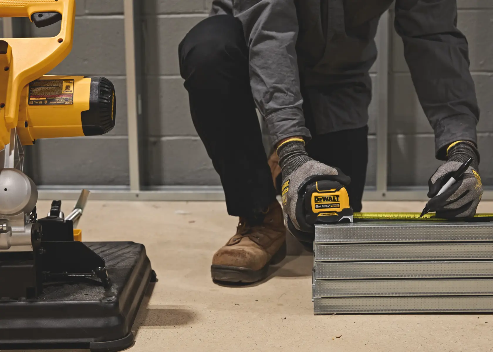 A person wearing gloves uses a DEWALT tape measure to measure metal studs, marking a measurement with a pen in a workshop setting. A part of a yellow power tool is visible on the left.
