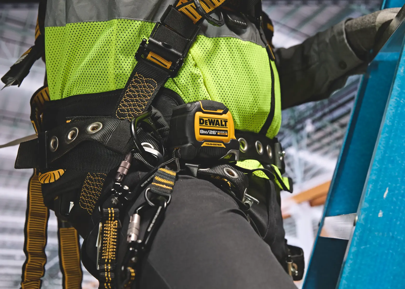 Close-up view of a DEWALT DWHT37926S tape measure attached to a worker's safety harness. The worker is wearing a fluorescent green safety vest and climbing a blue ladder in an indoor industrial setting.