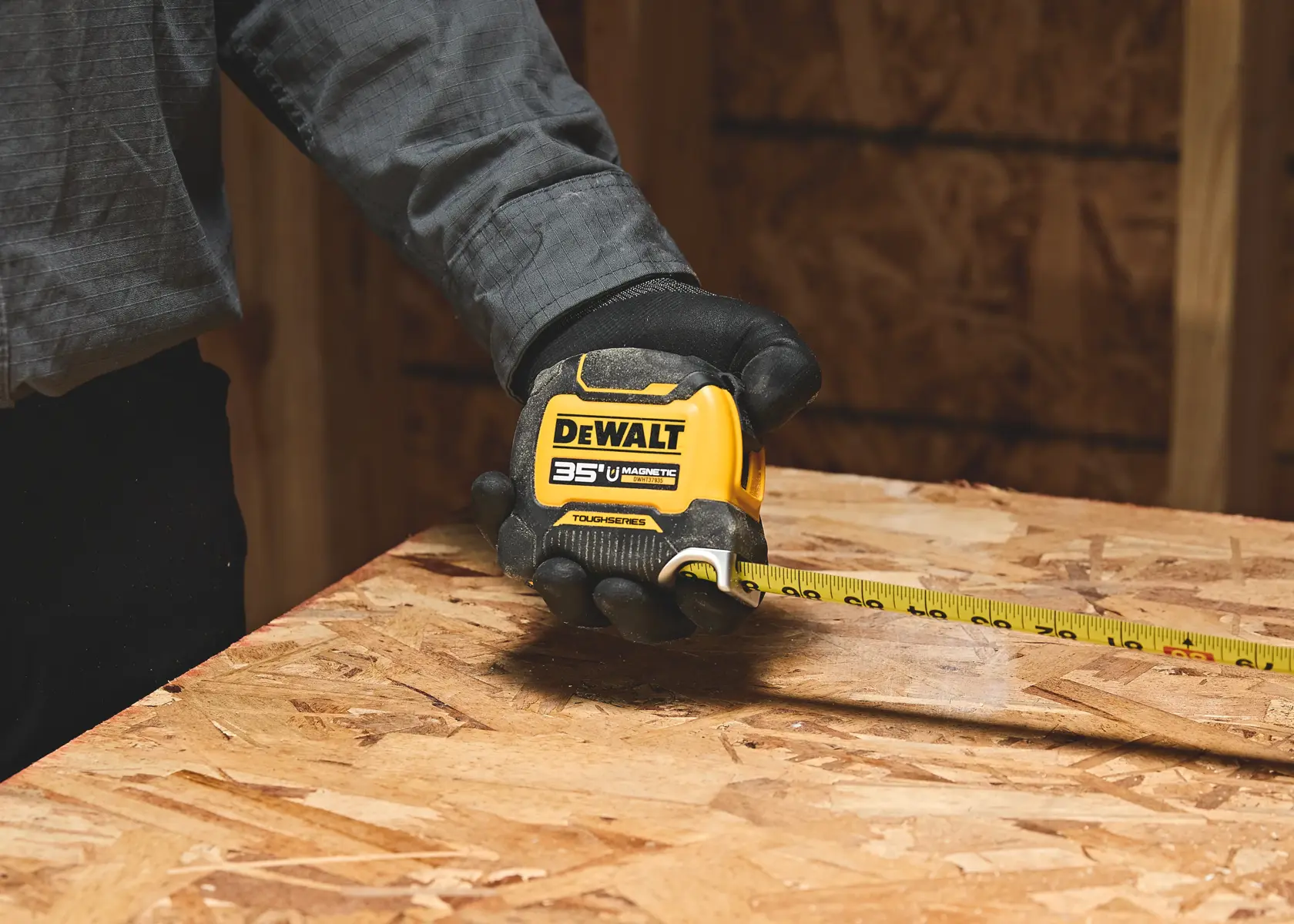 A person wearing gloves uses a yellow DEWALT 35 ft magnetic tape measure to measure a wooden board inside a workshop.