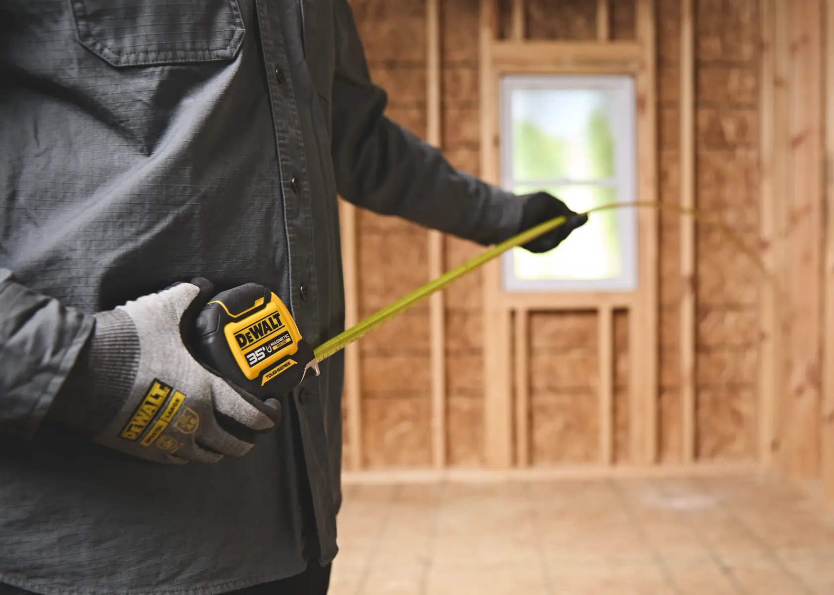 A person wearing gloves and a dark shirt is using a DEWALT 35-foot tape measure in a partially constructed wooden room.