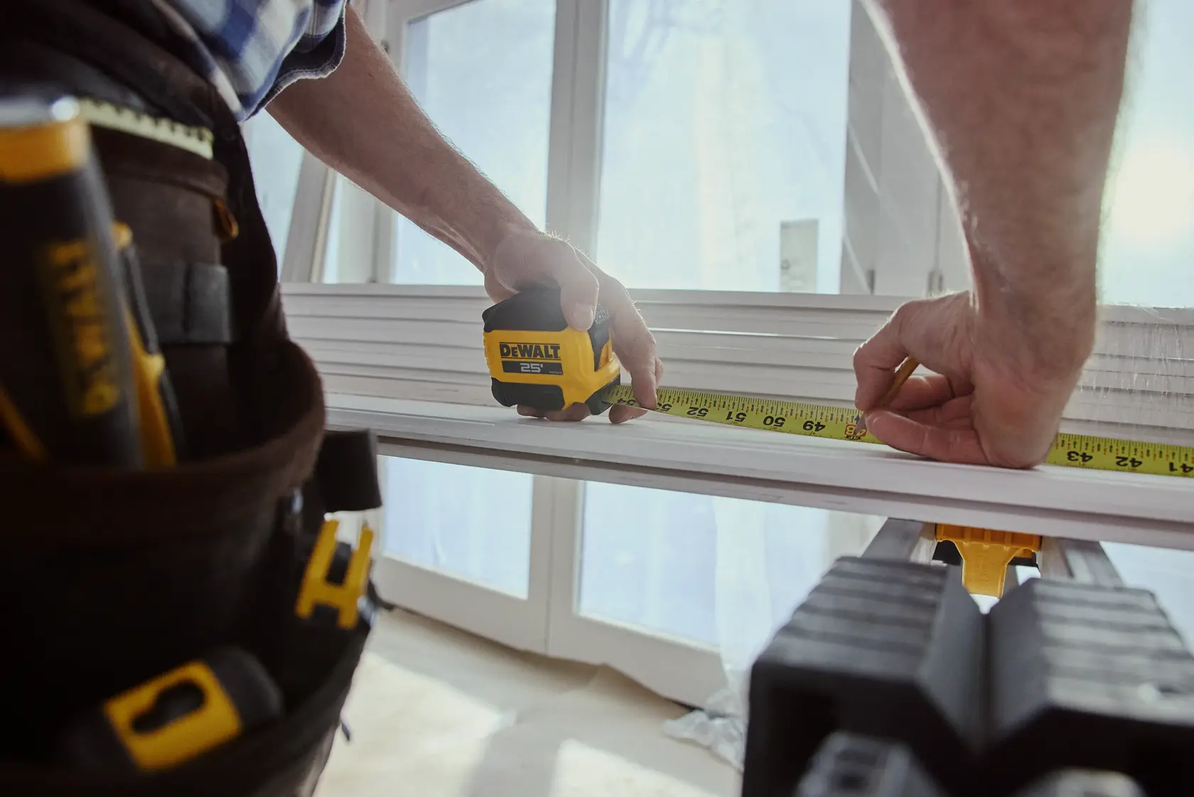 A person uses a DEWALT 25-foot tape measure to measure a piece of wood on a workbench. The person is wearing a tool belt and working indoors near large windows.
