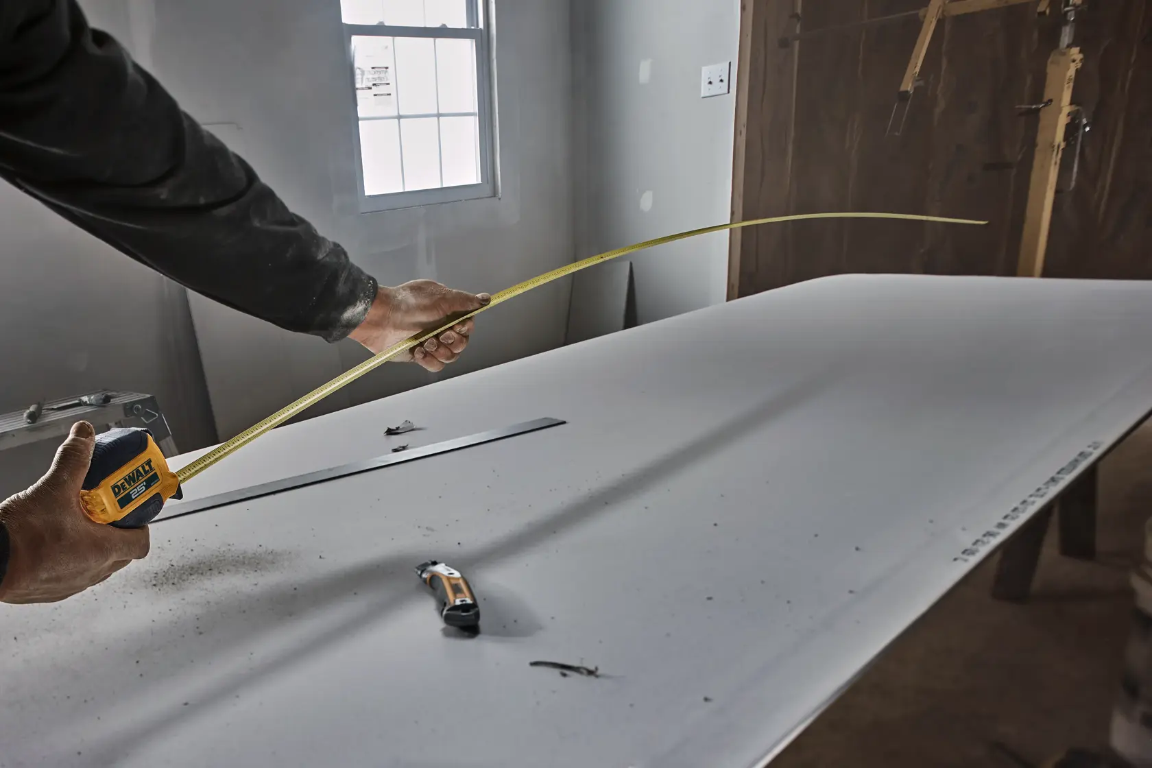 A person is using a DEWALT tape measure to measure a piece of drywall on a worktable, with a utility knife and a metal ruler also visible on the table.