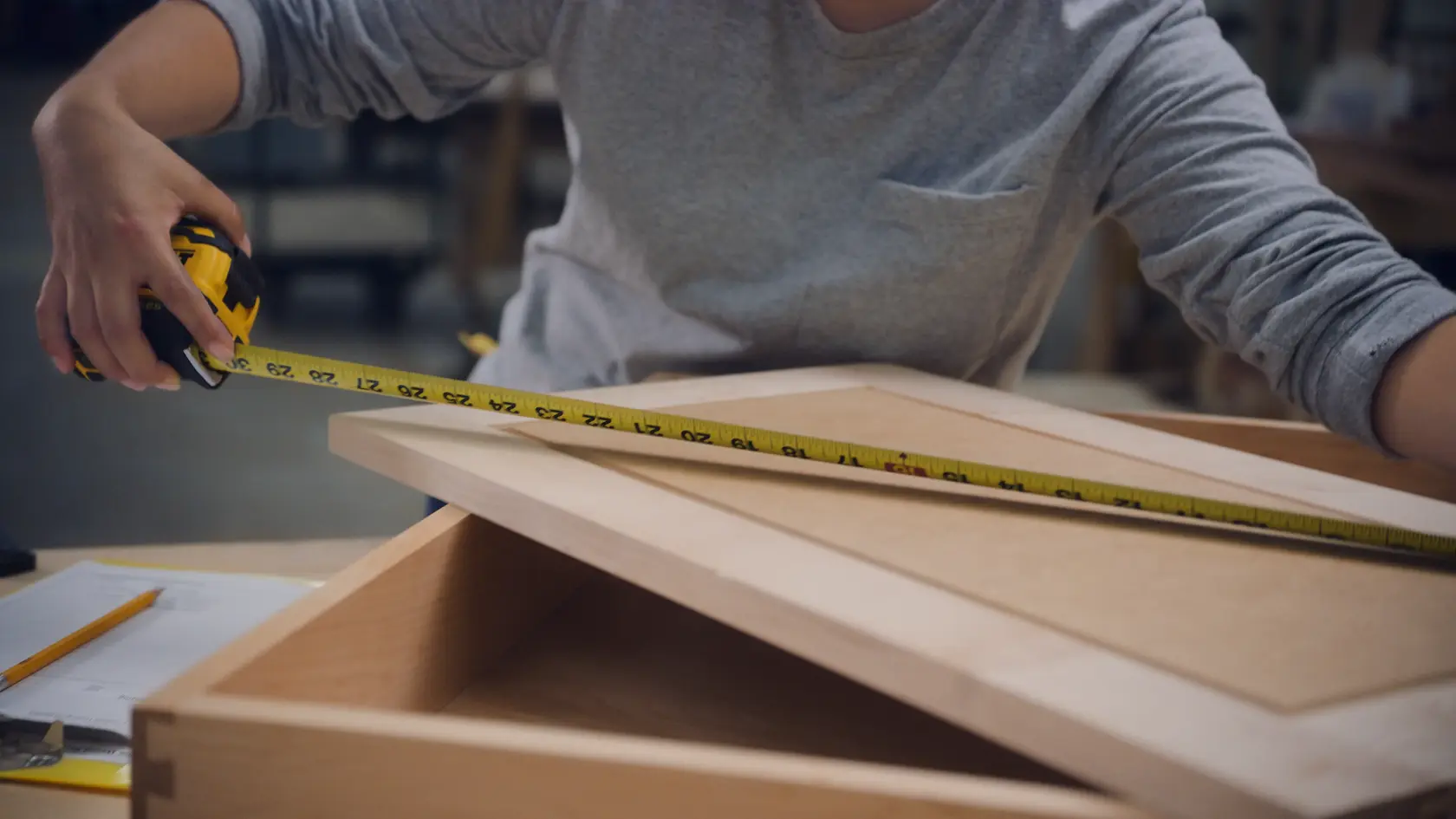 A person is using a yellow and black DEWALT tape measure to measure a wooden frame on a table in a workshop.