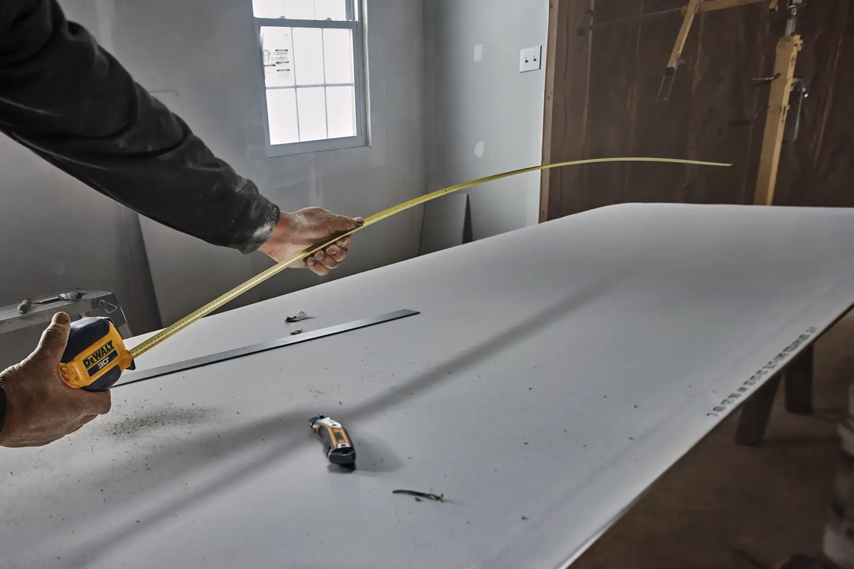 A person uses a DEWALT measuring tape to measure a piece of drywall in a construction setting. The tape is extended across the surface, and there are other tools visible on the drywall sheet.