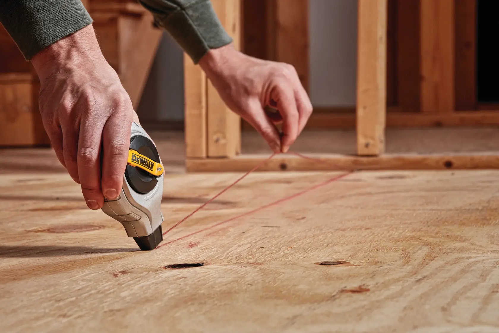 A person uses a DEWALT chalk line tool to mark a straight line on a plywood floor during construction.