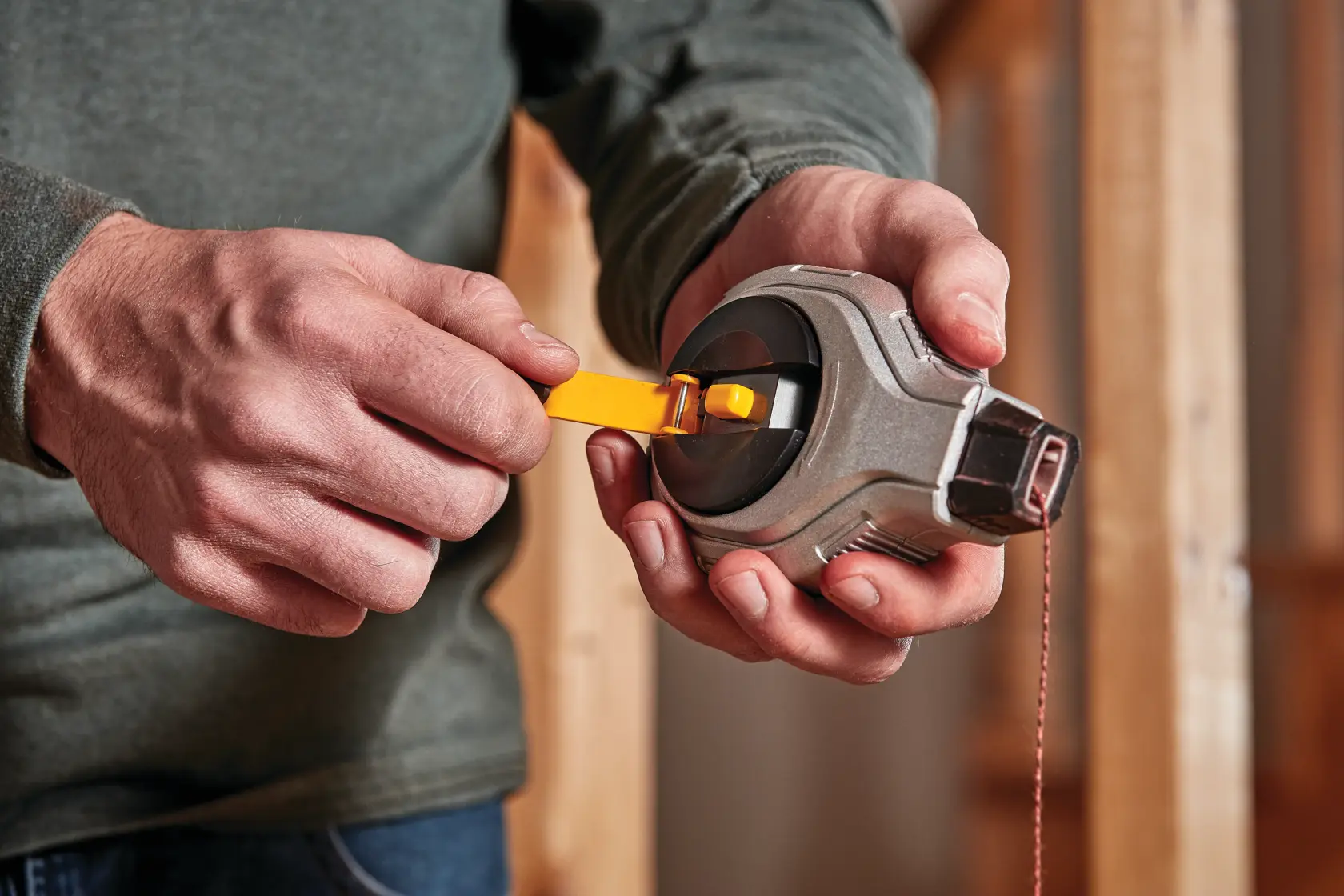 Close-up of a person’s hands using a DEWALT chalk reel tool, loading chalk powder with a yellow lever, against a background of unfinished wooden construction.