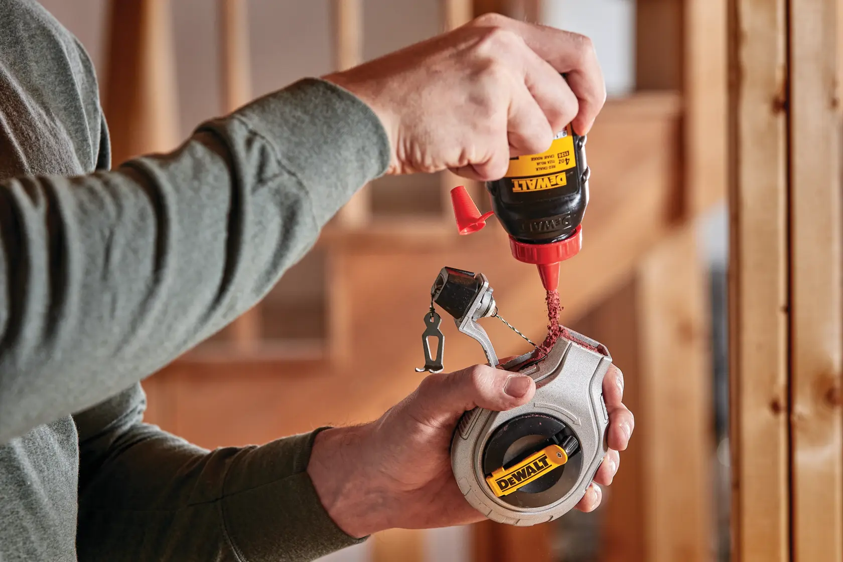 A person pours red chalk powder from a DEWALT bottle into a DEWALT chalk line tool, with a construction setting in the background.
