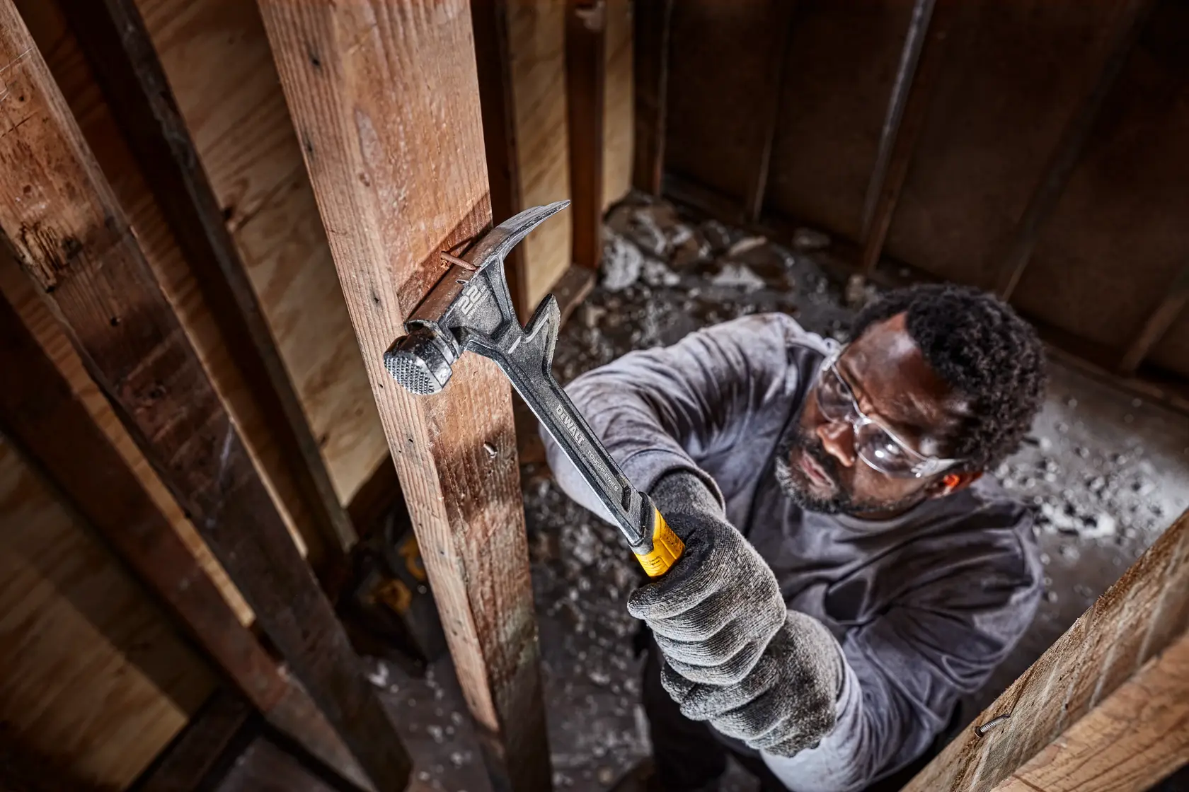 A person wearing gloves uses a DEWALT hammer to work on a wooden beam inside a construction site. The person's face is blurred for privacy.