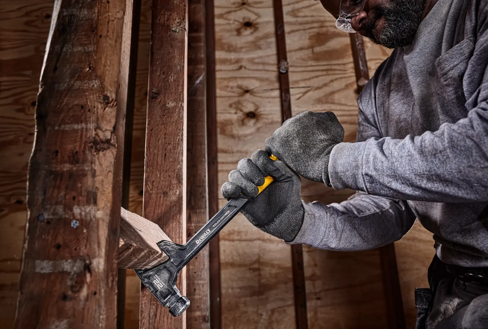 A person wearing safety gloves uses a DEWALT hammer to remove a wooden board from a construction frame inside a building.