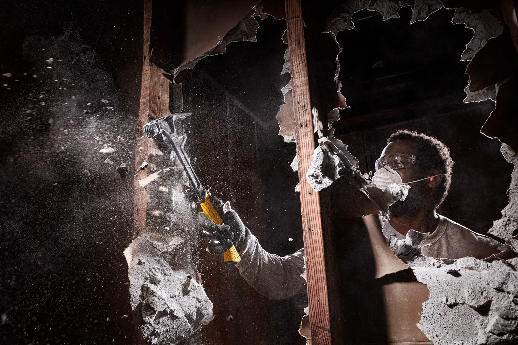 A person wearing gloves uses a yellow-handled DEWALT hammer to break a wall, causing debris and dust to fly in a construction or demolition setting. The person’s face is blurred for privacy.