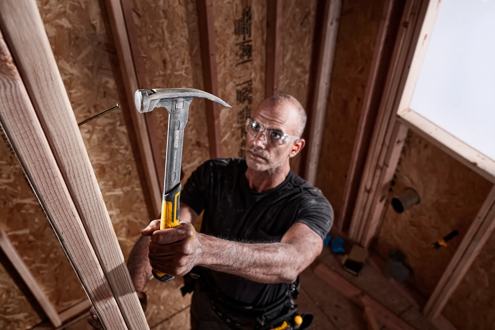 A person using a DEWALT hammer to drive a nail into a wooden beam inside a construction site. The hammer has a metal head and a yellow handle, and the person's face is blurred.