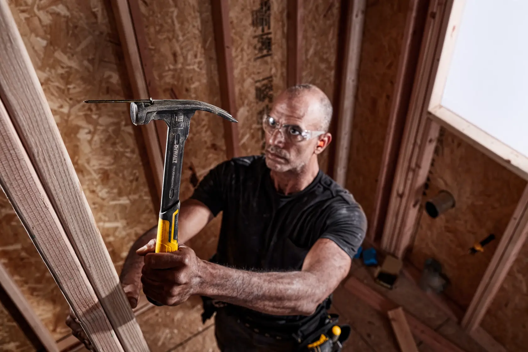 A person with a blurred face uses a DEWALT hammer to work on wooden framing inside a construction site. The hammer has a yellow and black handle and is being held firmly.