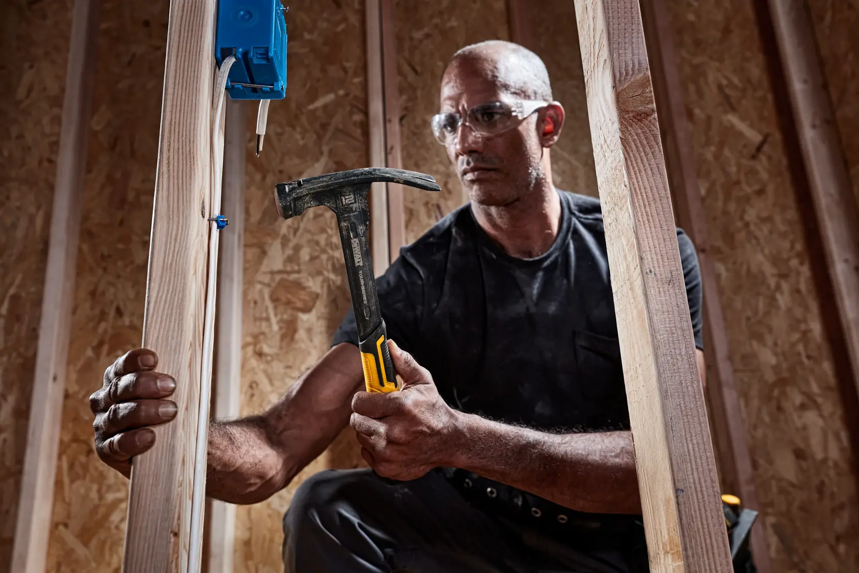 A person using a DEWALT hammer (model DWHT51135X) to work on wooden framing in a construction setting. The person is holding the hammer with a yellow and black handle near electrical wiring and a blue electrical box.