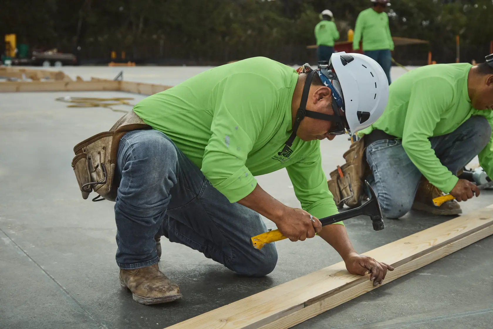 A worker wearing a helmet and green shirt kneels on a construction site, using a DEWALT hammer to secure a piece of wood. Other workers in green shirts are visible in the background.
