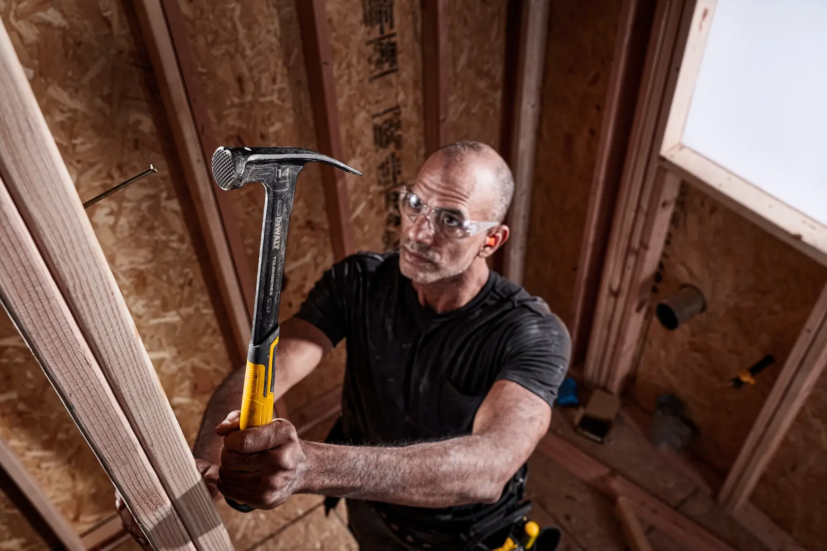 A person with a blurred face is using a DEWALT claw hammer with a yellow handle to drive a nail into wooden framing inside a construction site.