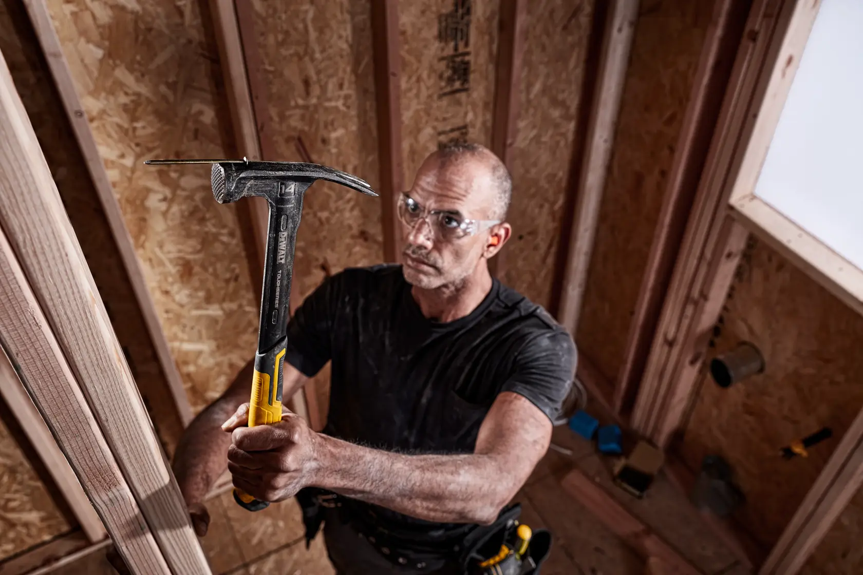 A person with a blurred face is holding a DEWALT hammer (DWHT51138X) with a yellow handle inside a wooden structure under construction. The setting suggests a work site or construction environment.