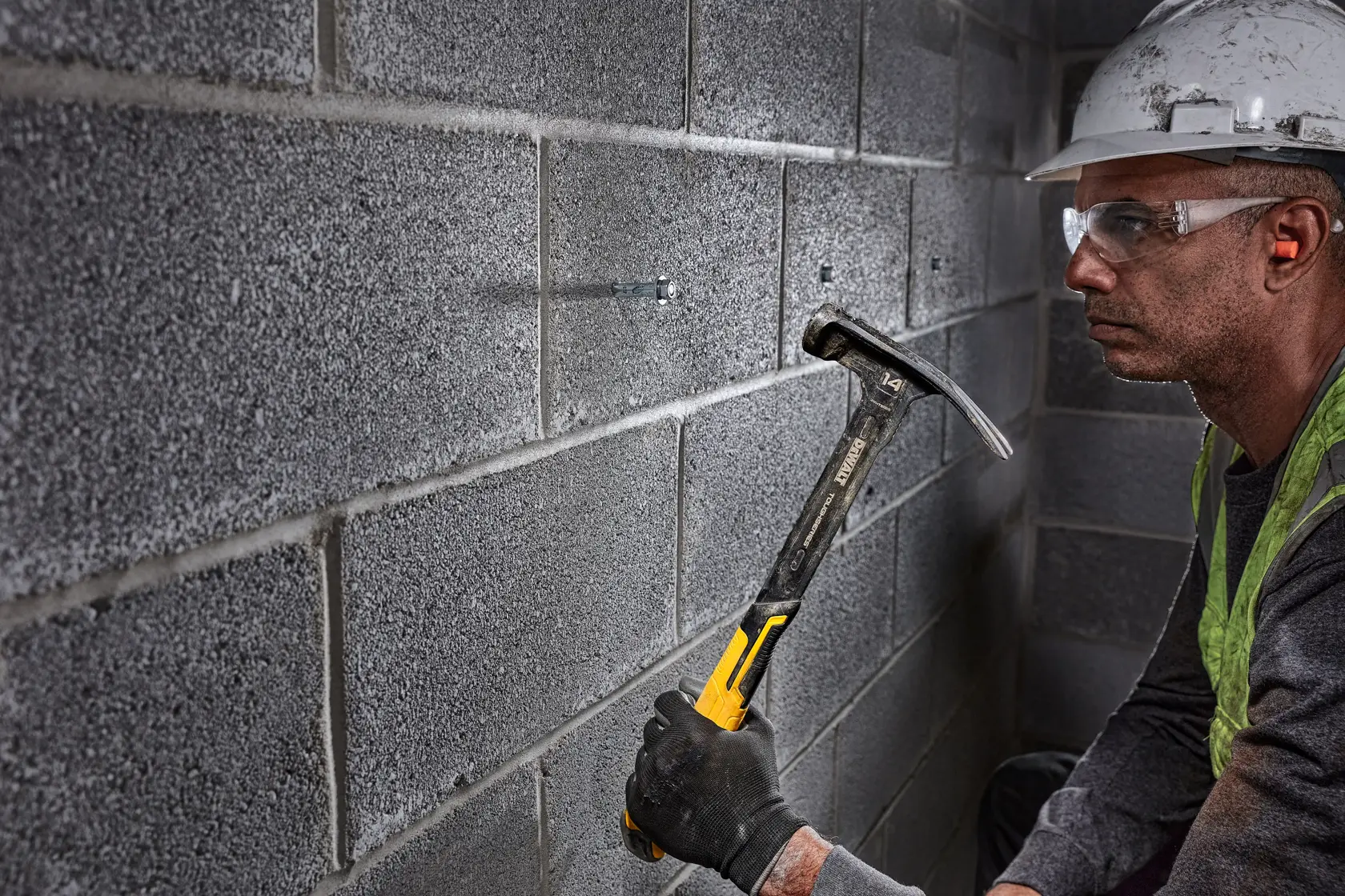 A person wearing a safety helmet and gloves uses a DEWALT claw hammer to drive a fastener into a concrete block wall.