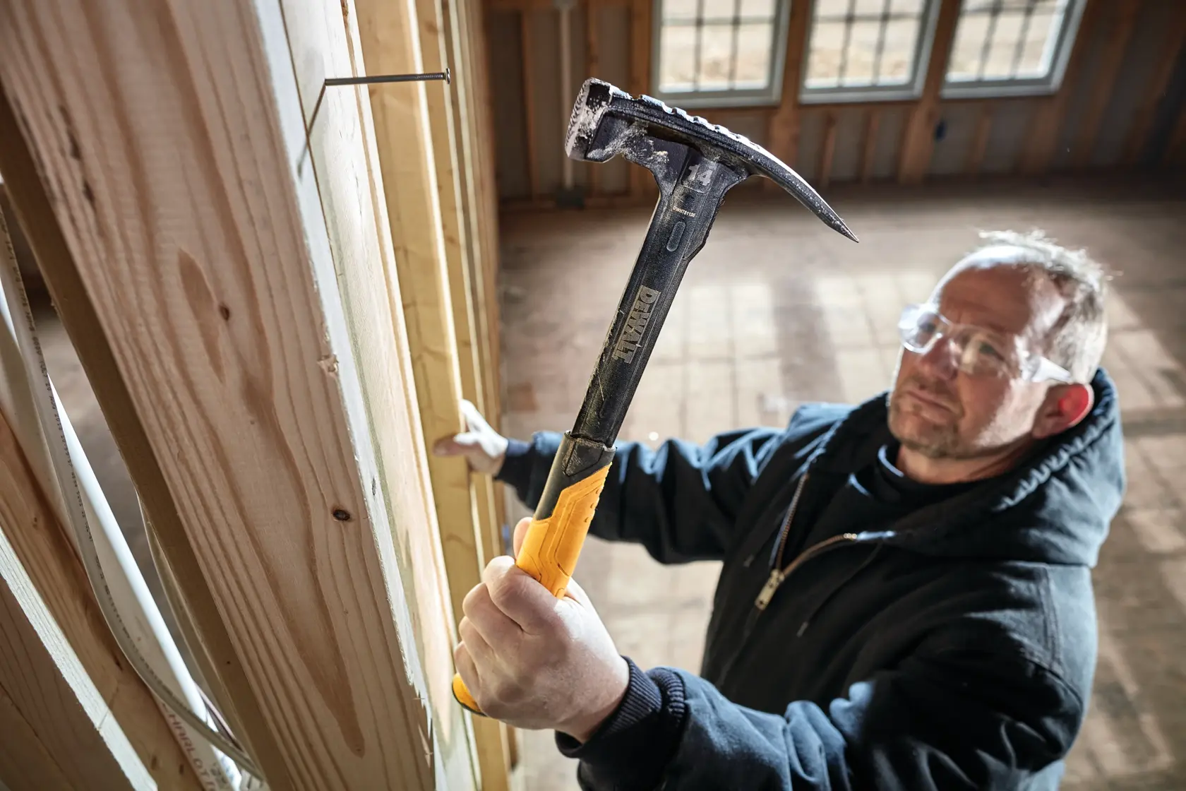 A person using a DEWALT hammer with a yellow handle to drive a nail into a wooden beam inside a building under construction. The face of the person is blurred for privacy.