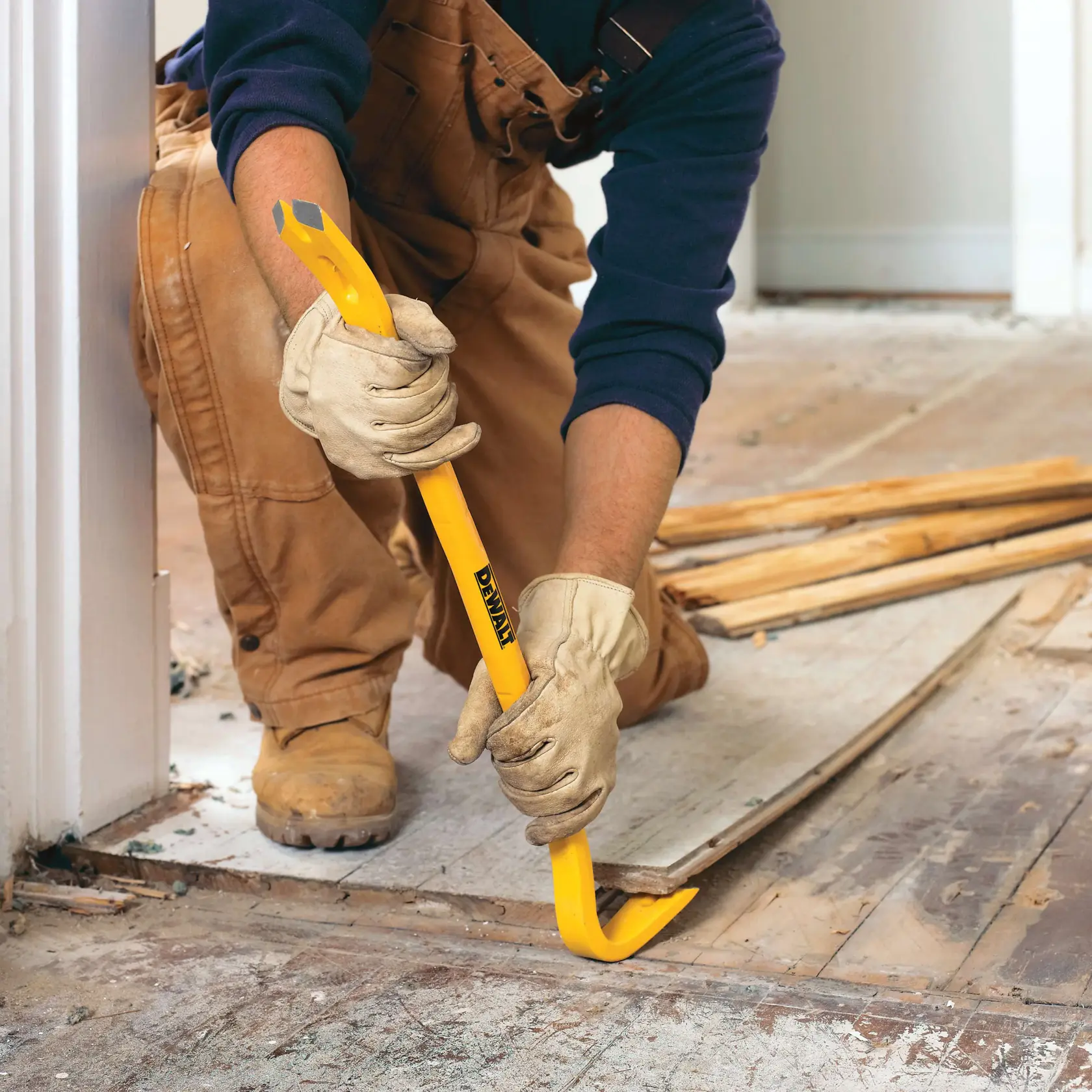 A person wearing work gloves, brown overalls, and boots uses a yellow DEWALT pry bar to remove wooden boards from a floor during construction or demolition.