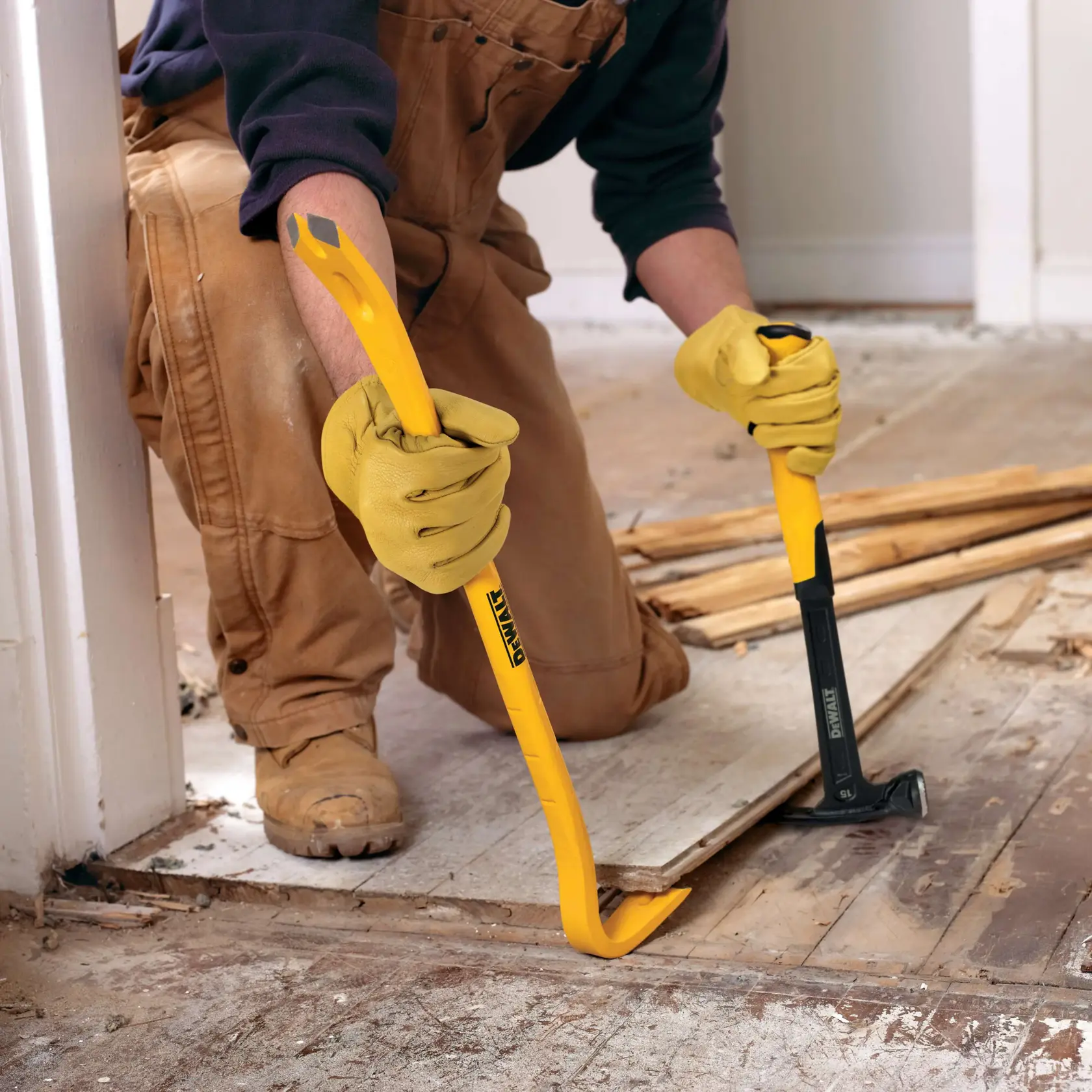 A person wearing gloves and work overalls uses a yellow DEWALT pry bar and a black DEWALT hammer to remove wooden flooring.
