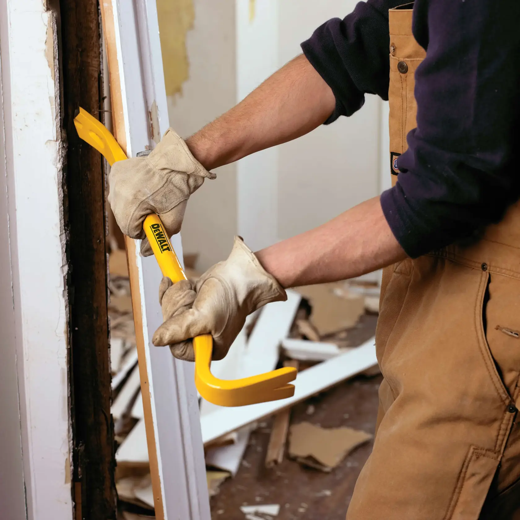 A person wearing gloves and work clothes uses a yellow DEWALT pry bar to remove trim from a doorway during demolition work.