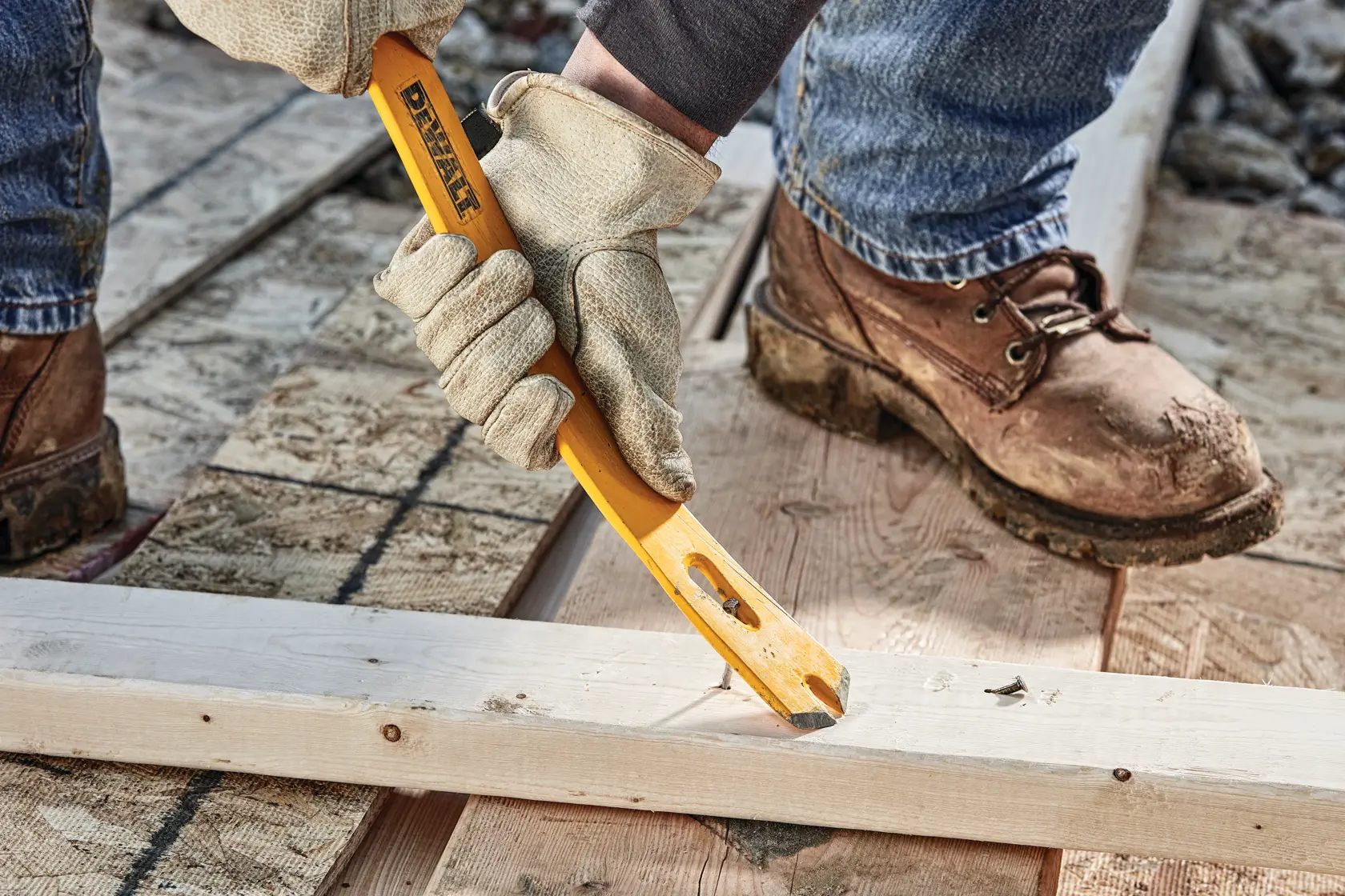A person wearing work gloves and boots uses a DEWALT yellow pry bar to remove nails from a wooden plank on a construction site.