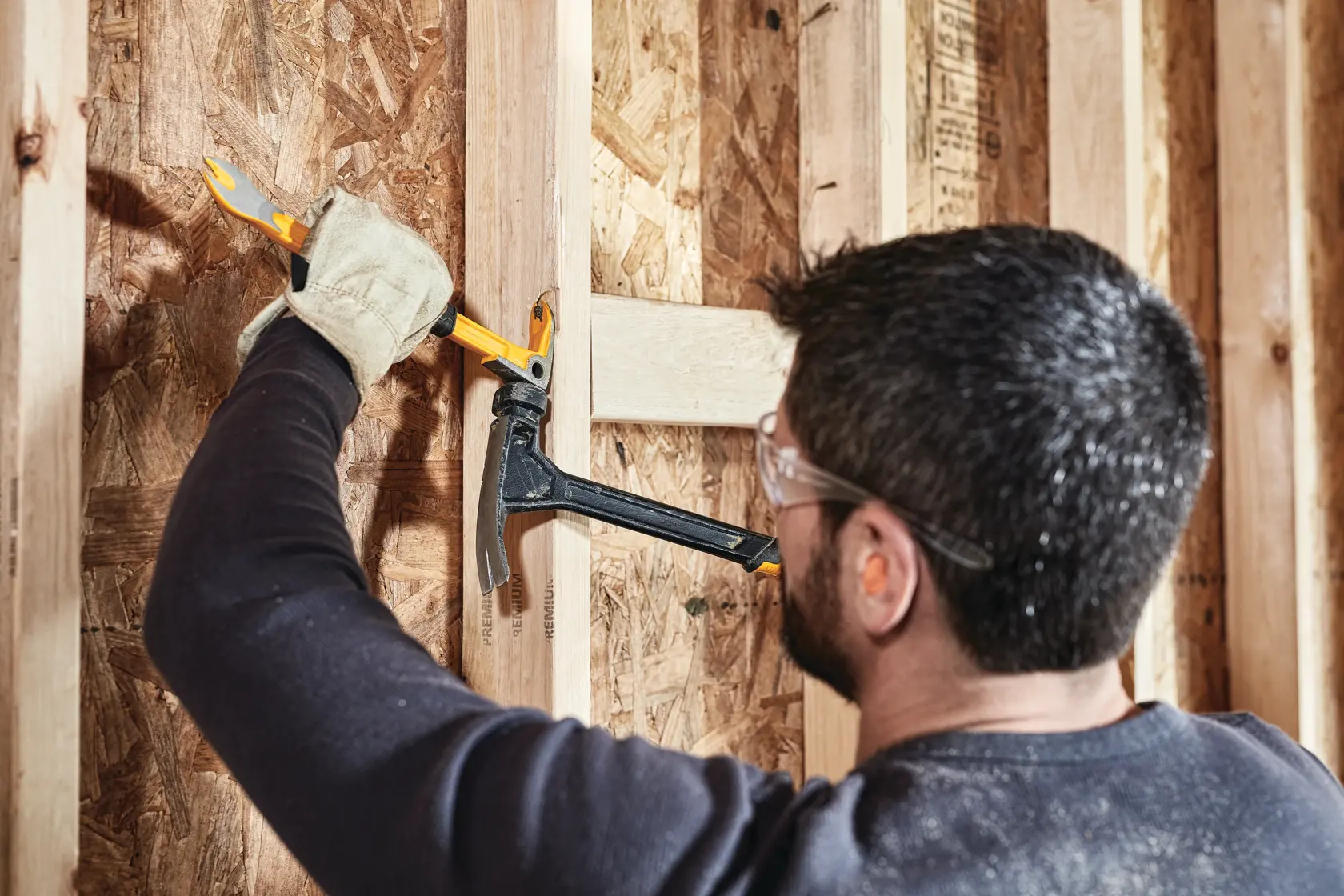 A person wearing safety goggles and gloves uses a DEWALT demolition tool to remove wooden planks from a wall during construction.