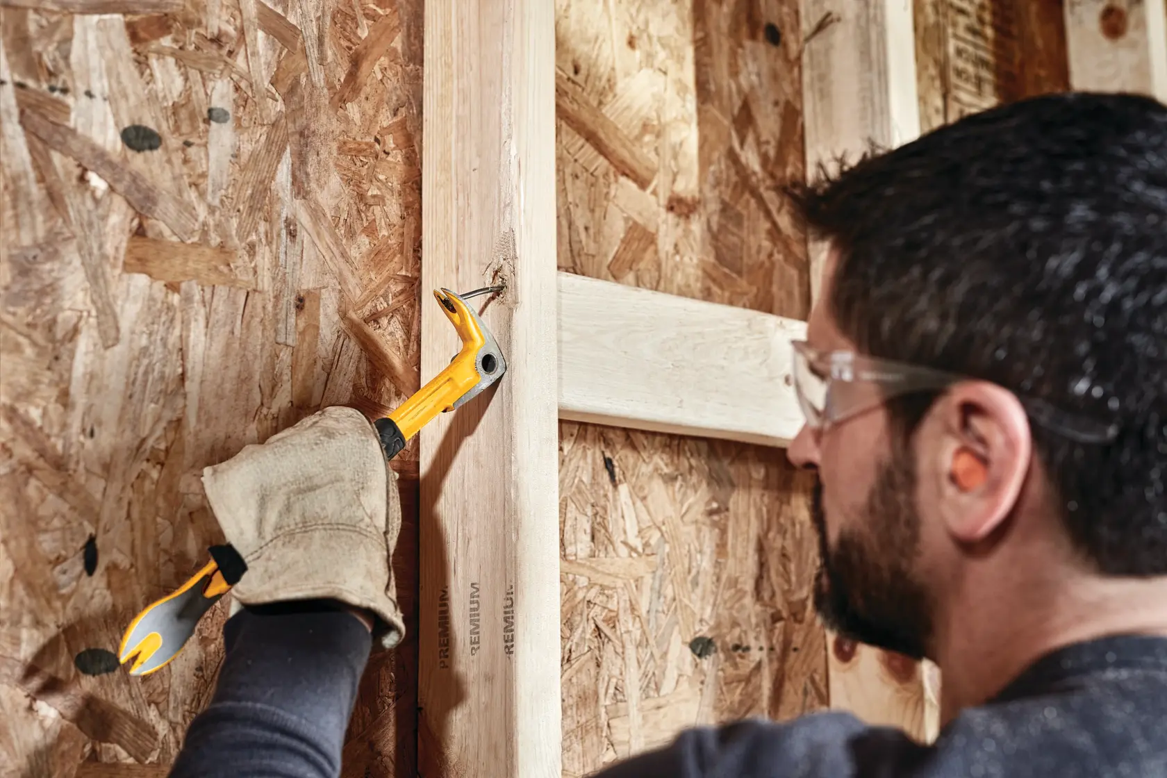 A person with gloves uses a DEWALT yellow hand tool, specifically a claw bar, to remove a nail from a wooden stud during construction. The person's face is blurred for privacy.