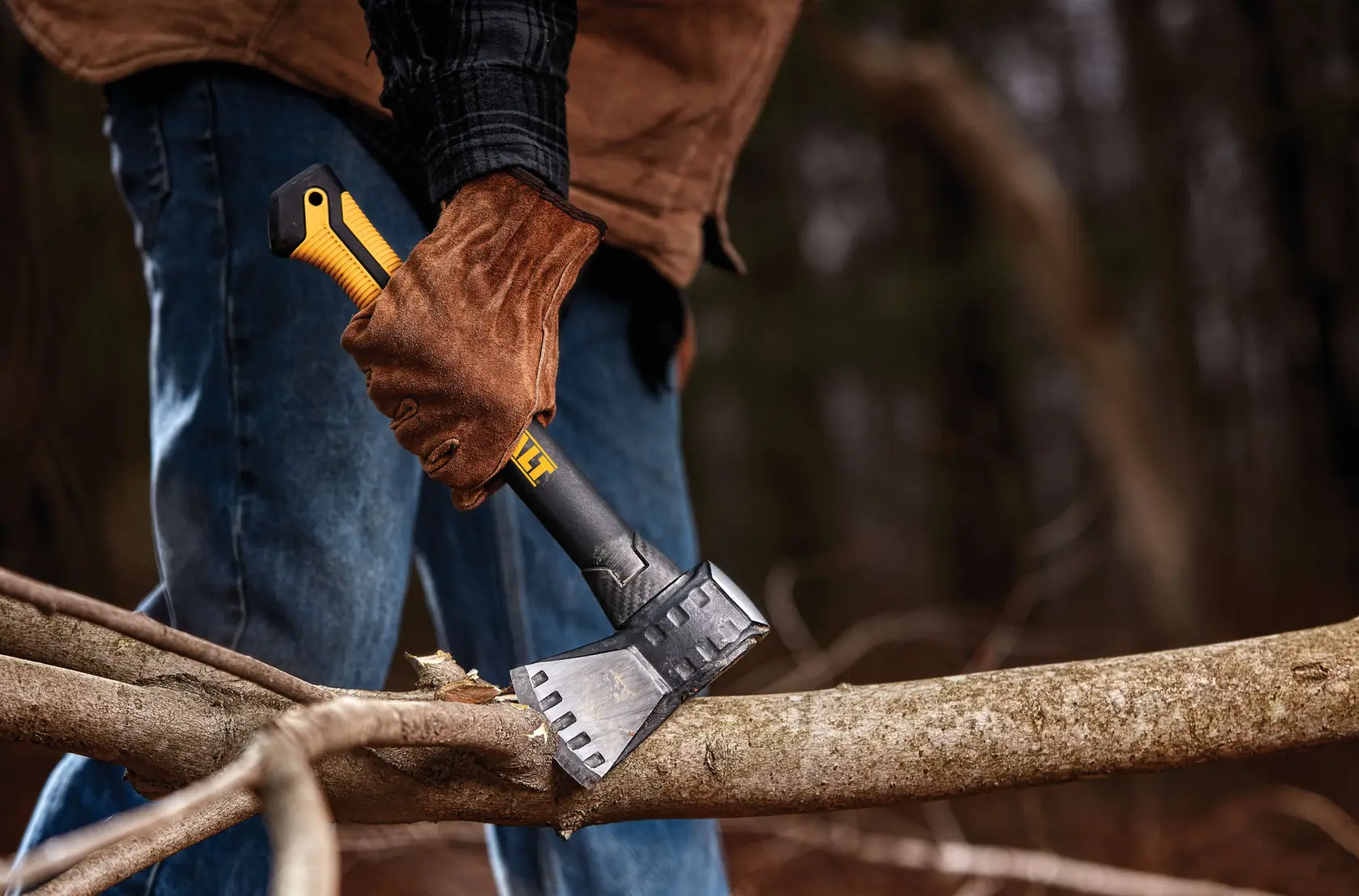 A person wearing brown gloves and a jacket is using a DEWALT axe to chop a thick branch outdoors.