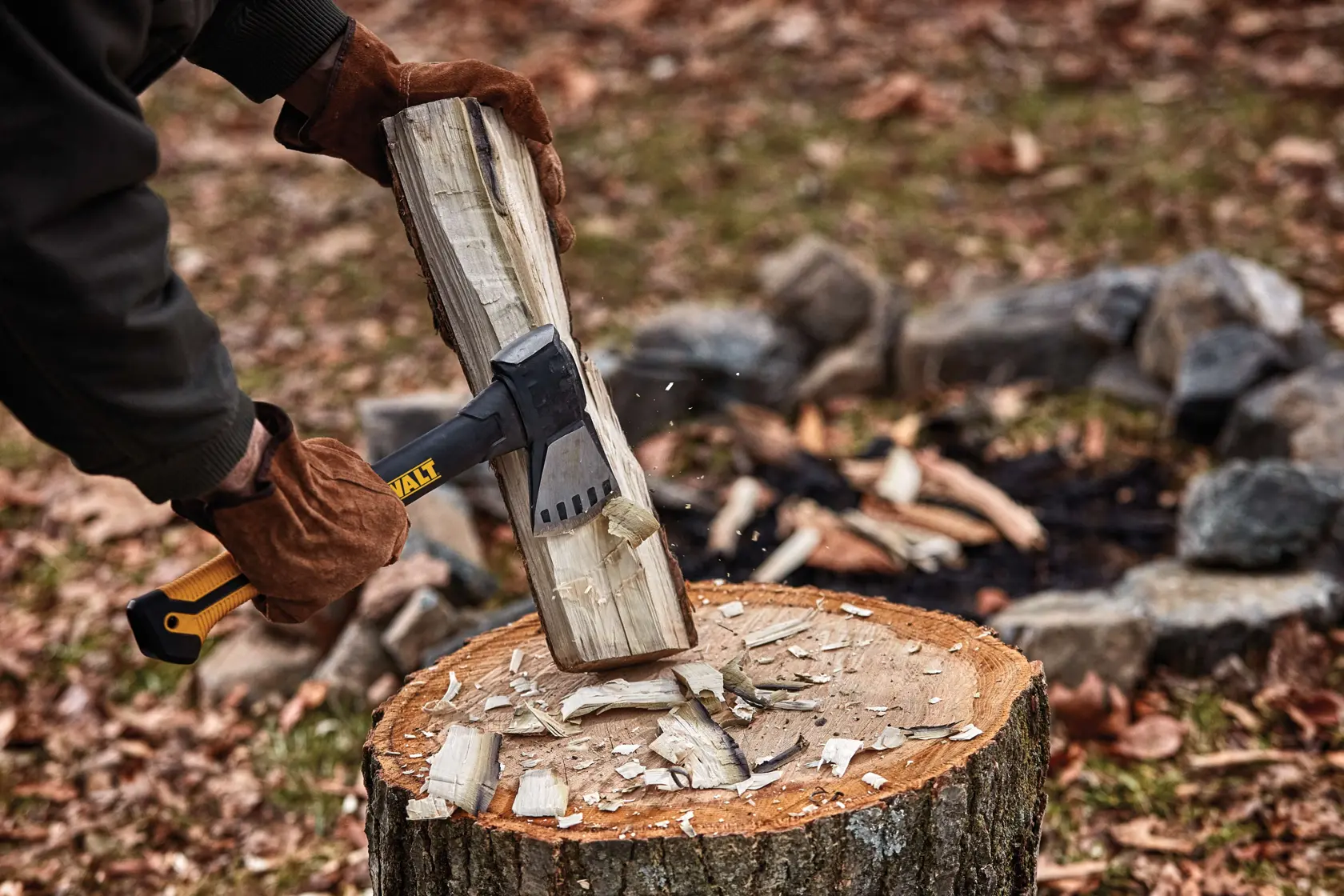A person wearing brown gloves uses a DEWALT-branded axe to split a piece of wood on a tree stump outdoors, with wood chips scattered and a stone fire pit in the background.