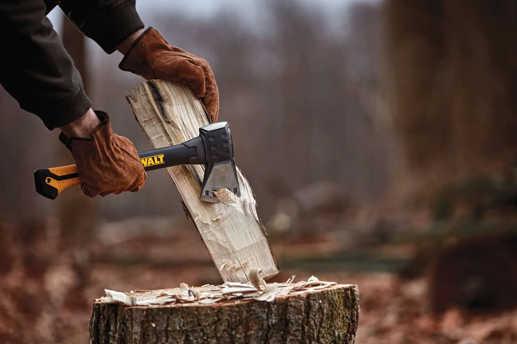 A person wearing brown gloves uses a DEWALT axe to split a piece of wood on a tree stump outdoors.