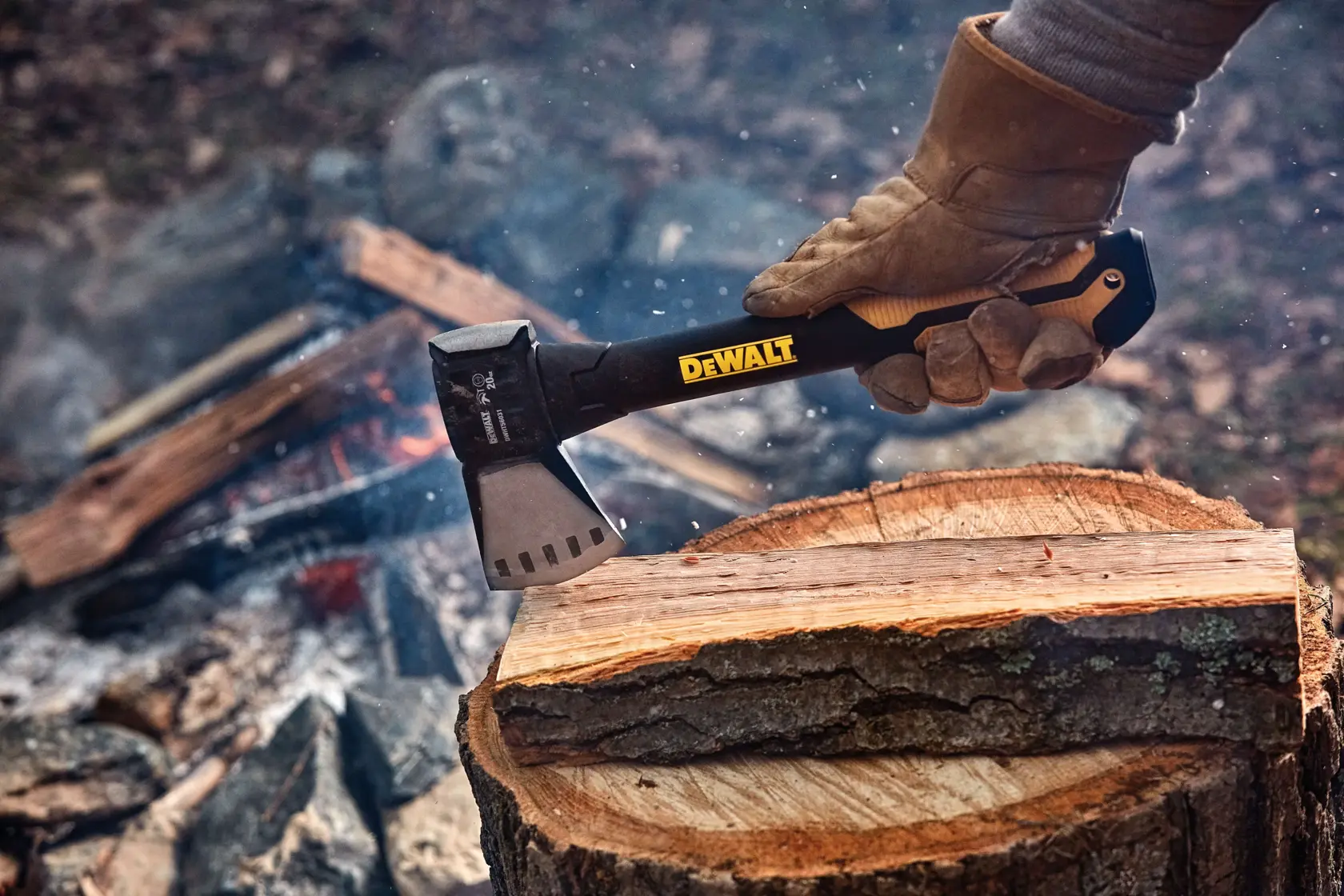A person wearing gloves uses a DEWALT hatchet to split a piece of wood outdoors, with a campfire and other logs in the background.