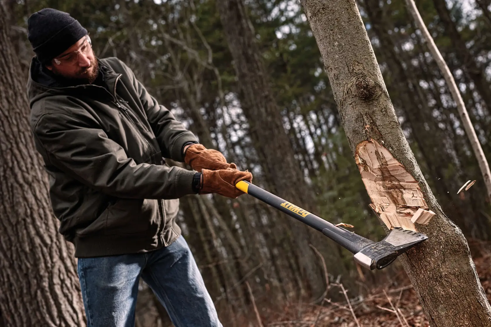 A person using a DEWALT axe (model DWHT56032) to chop a tree outdoors, wearing gloves and jacket with the face blurred for privacy. The axe is striking the tree, and wood chips are flying off.