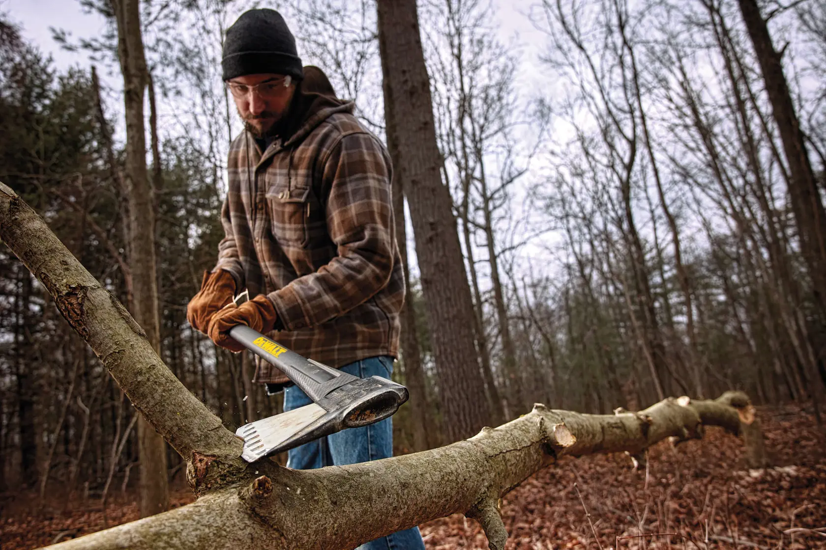 A person wearing gloves and a plaid jacket uses a DEWALT axe to chop branches from a fallen tree in a wooded area. The person's face is blurred.