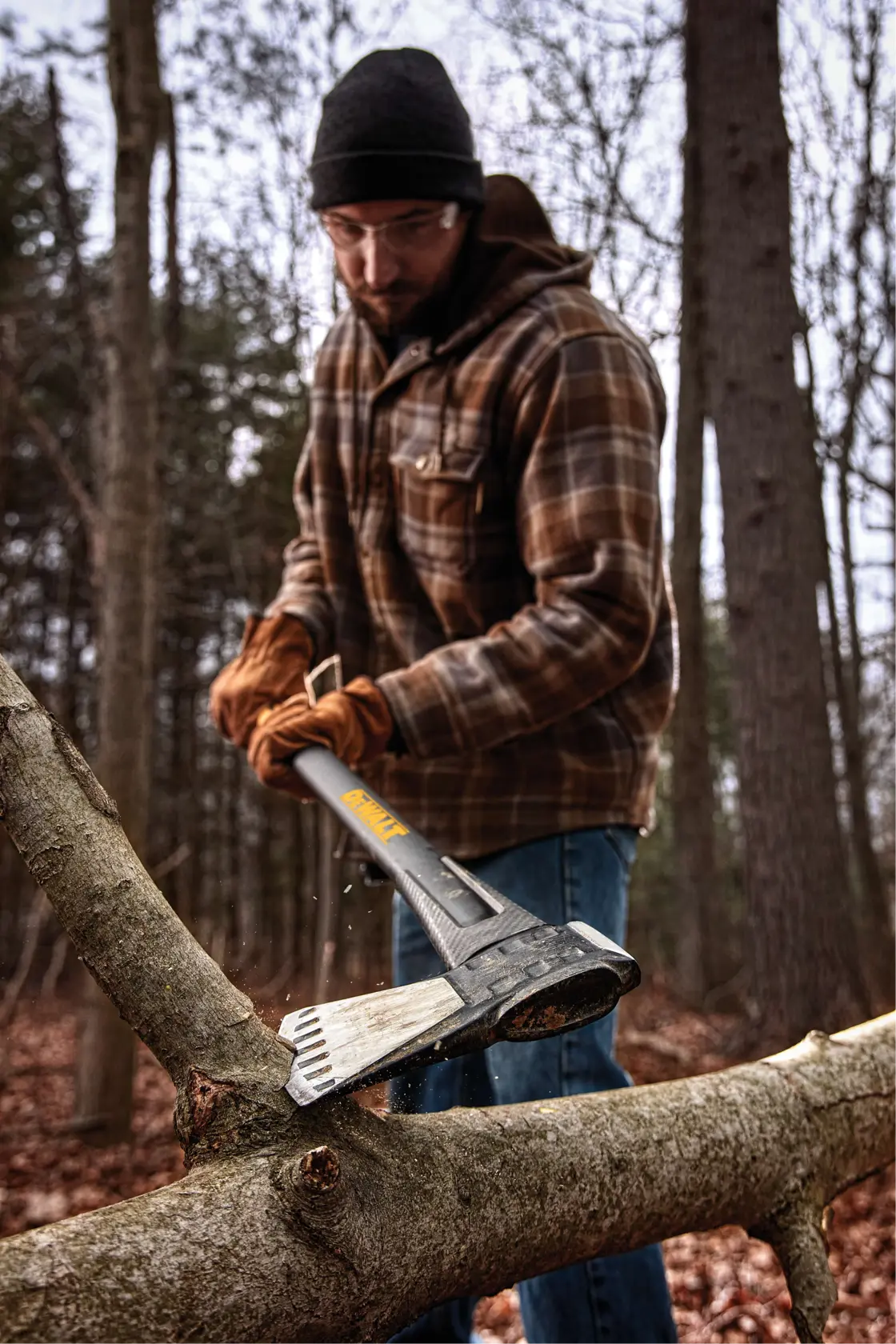 A person wearing a plaid jacket and gloves uses a DEWALT axe to chop a large tree branch outdoors in a wooded area. The axe blade is making contact with the branch, and wood chips are flying.