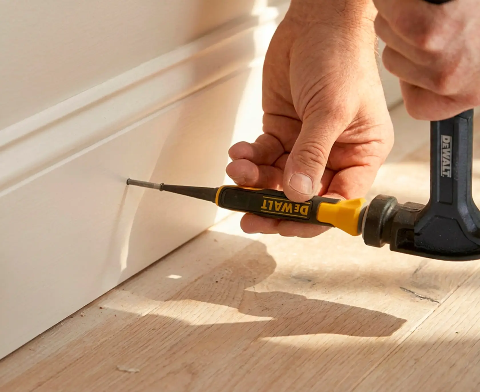 Hand uses DEWALT nail set tool and hammer on wood trim near floor.