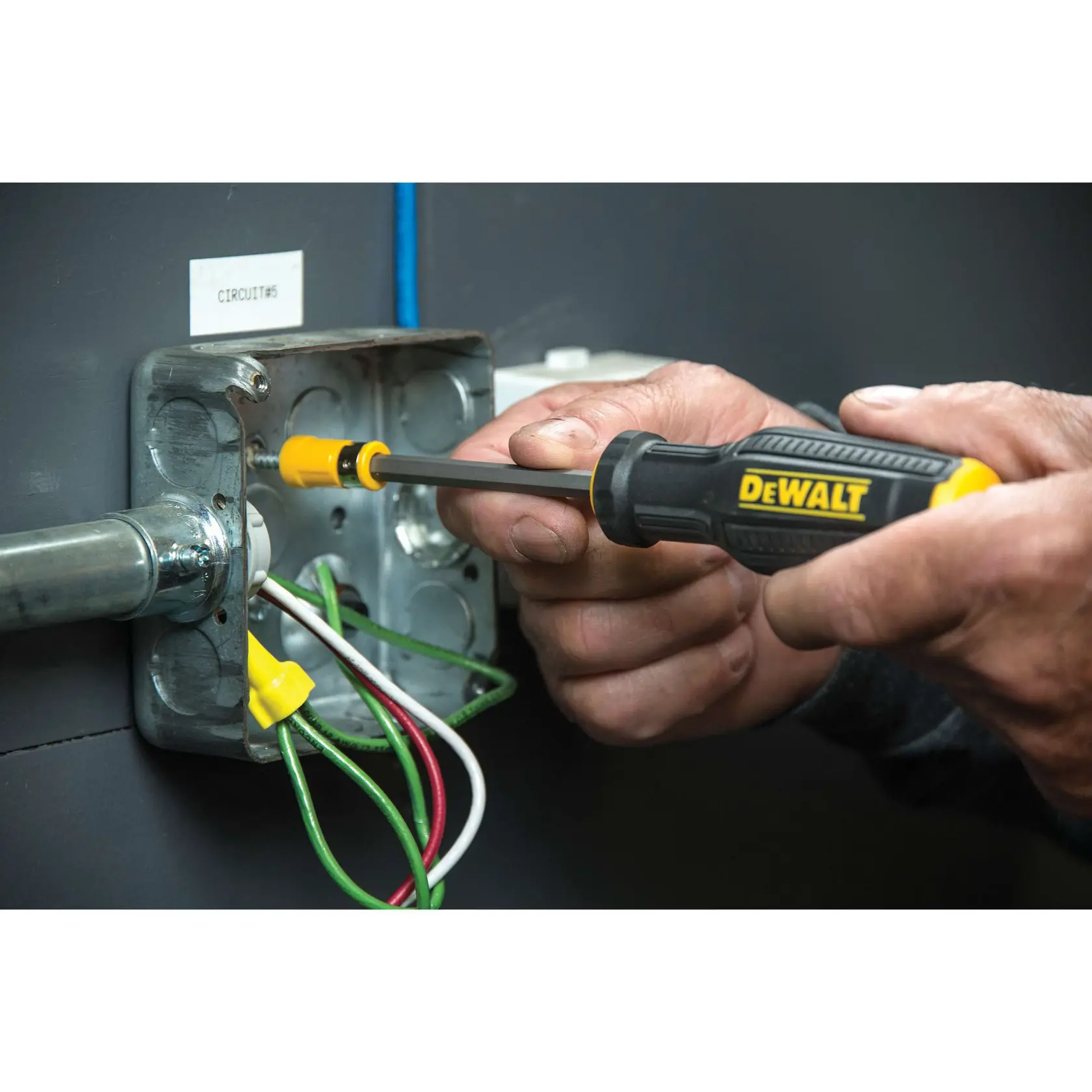 A person uses a DEWALT screwdriver to tighten a screw in an electrical outlet box with visible green, white, and red wires.