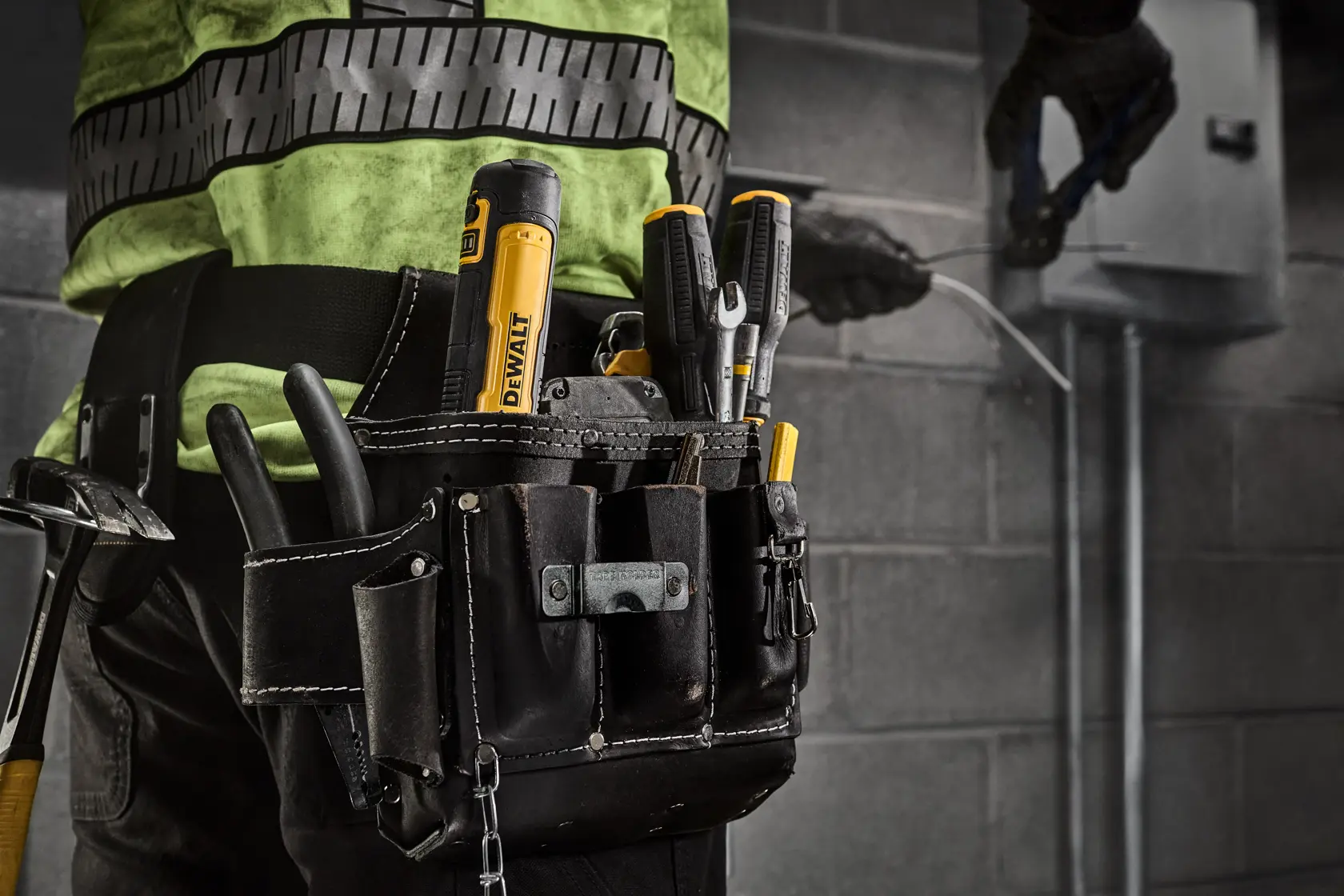 Close-up of a worker wearing a tool belt filled with various DEWALT hand tools, including screwdrivers, pliers, and a utility knife. The worker is performing maintenance work near an electrical box against a gray brick wall.