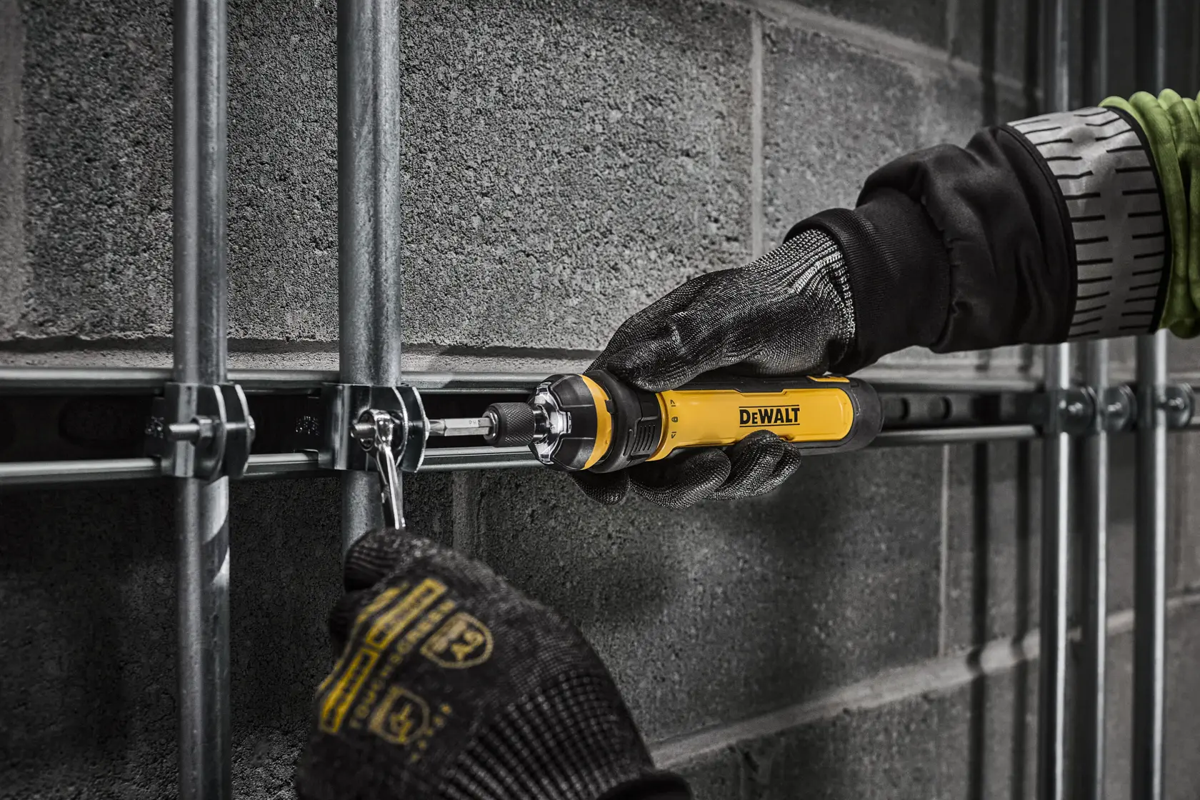 A person wearing protective gloves uses a yellow DEWALT screwdriver to tighten or loosen a bolt on a metal conduit mounted to a concrete wall.
