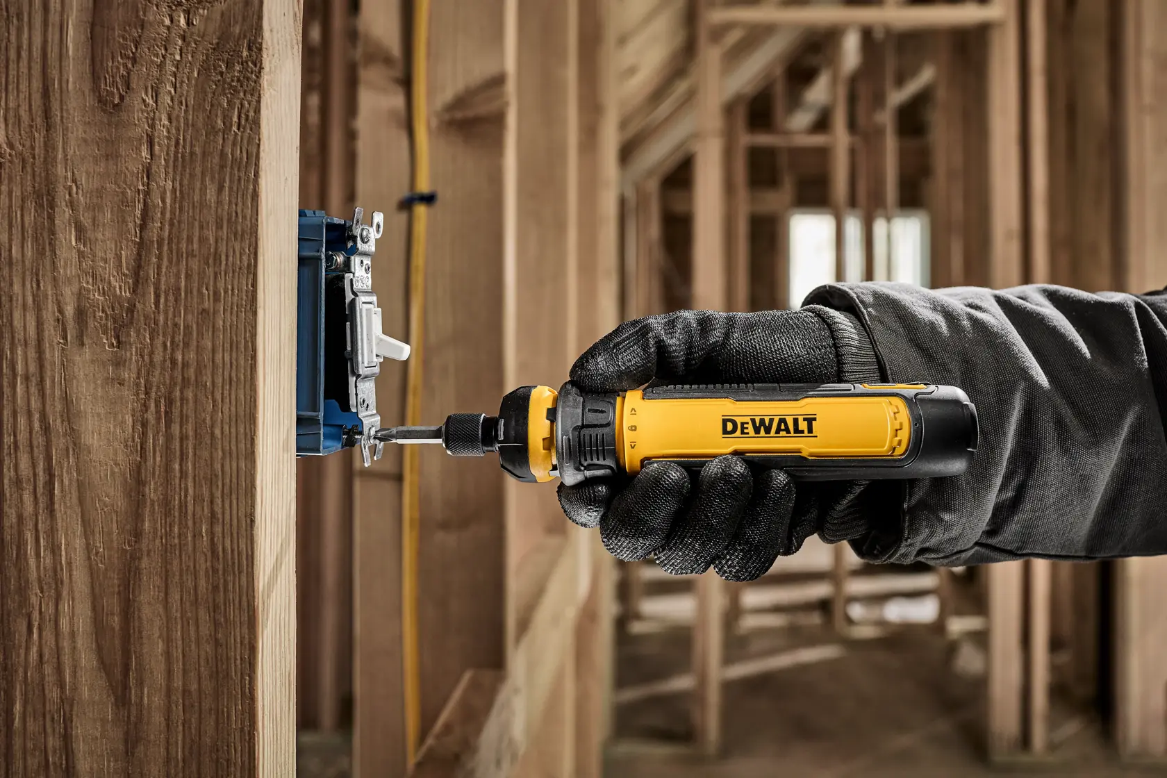 A gloved hand using a yellow and black DEWALT screwdriver to install or adjust an electrical outlet box on a wooden wall frame at a construction site.