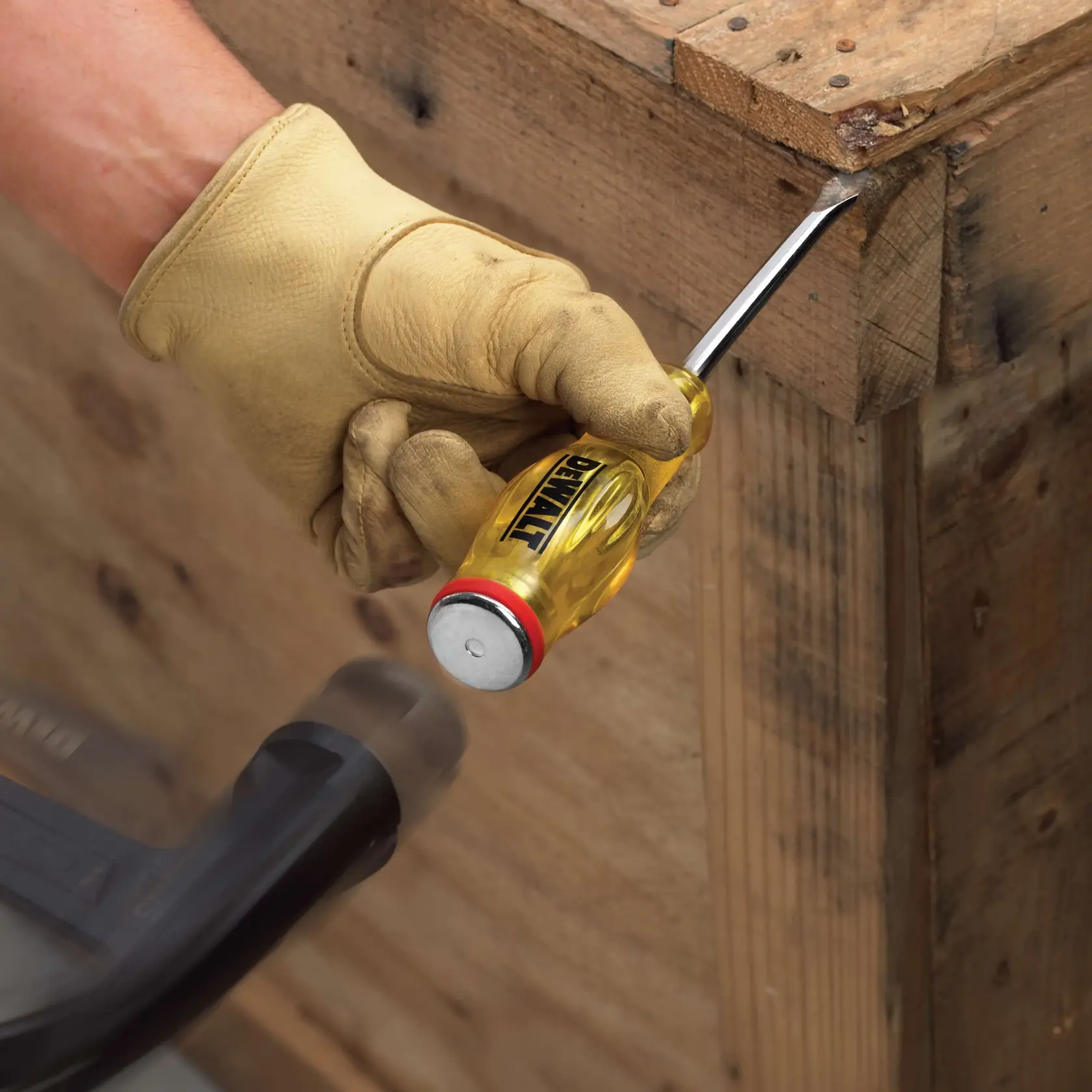 A gloved hand using a DEWALT flat head screwdriver with a yellow handle to pry apart wooden boards, while another tool strikes the screwdriver's metal end.