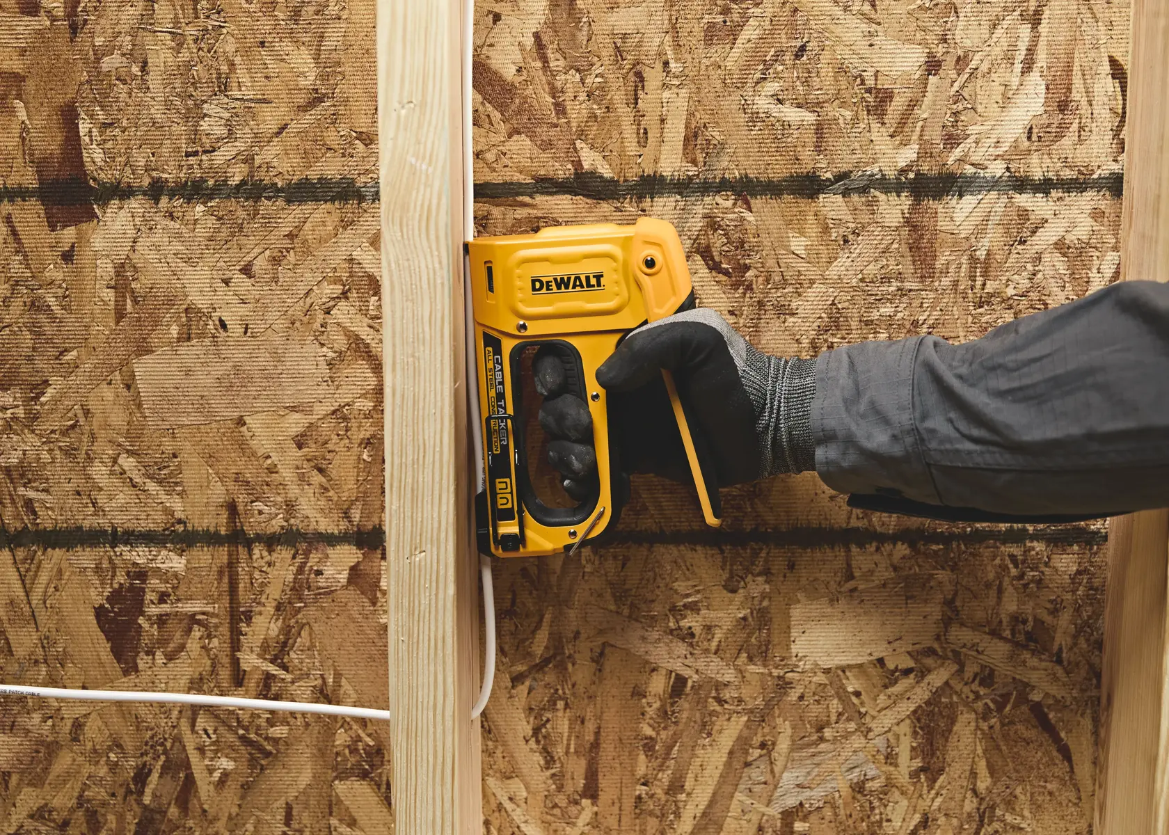 A person wearing a glove is using a yellow DEWALT staple gun on a wooden stud of a wall under construction, with plywood sheathing in the background.